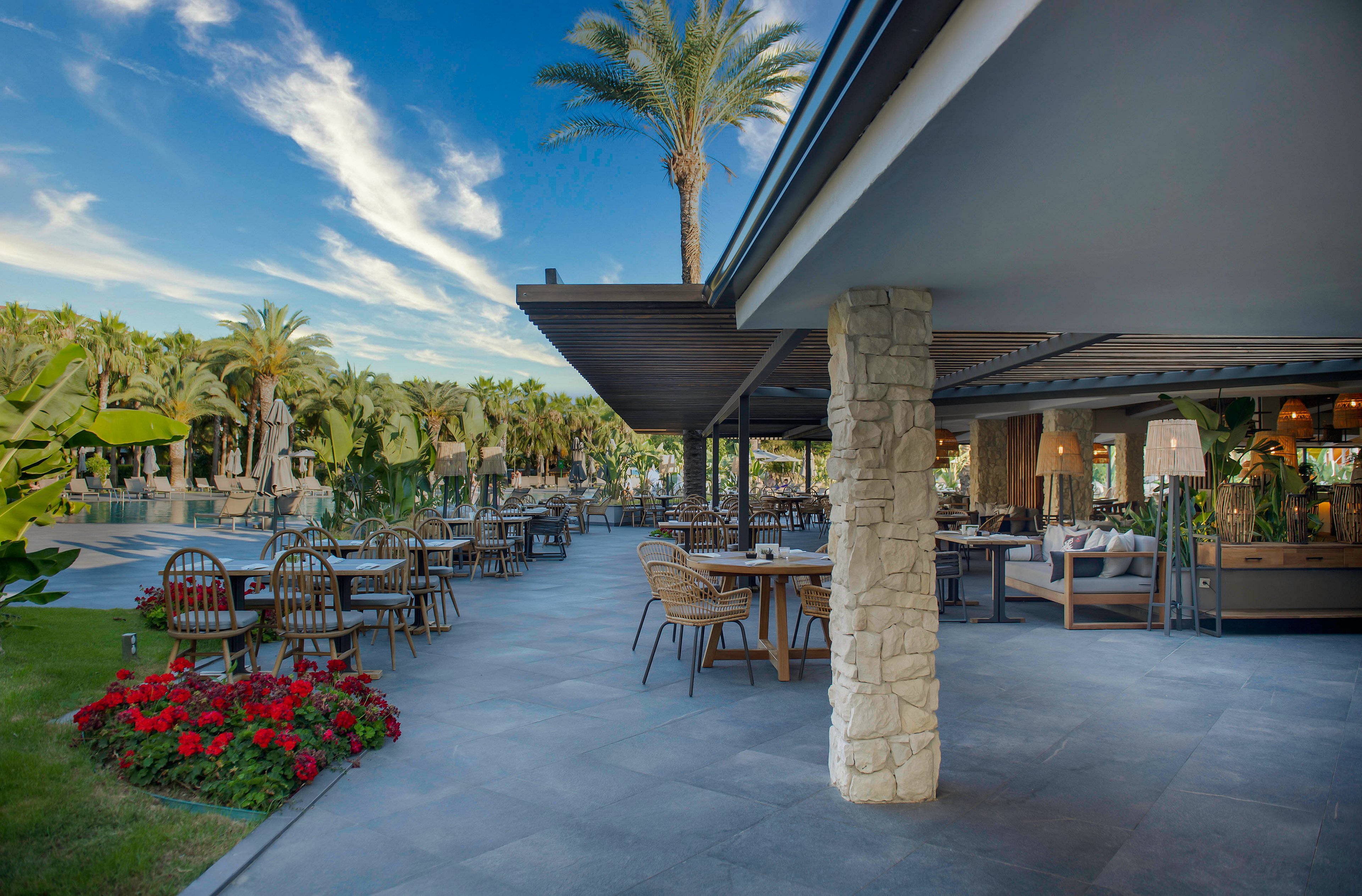 An open-air restaurant terrace is set with wooden tables and chairs beside a lush resort garden. Palm trees and tropical plants surround the seating area, creating a vacation atmosphere. The modern structure features stone columns, a shaded roof, and warm ambient lighting inside. The scene is calm and inviting under a bright blue sky with scattered clouds.