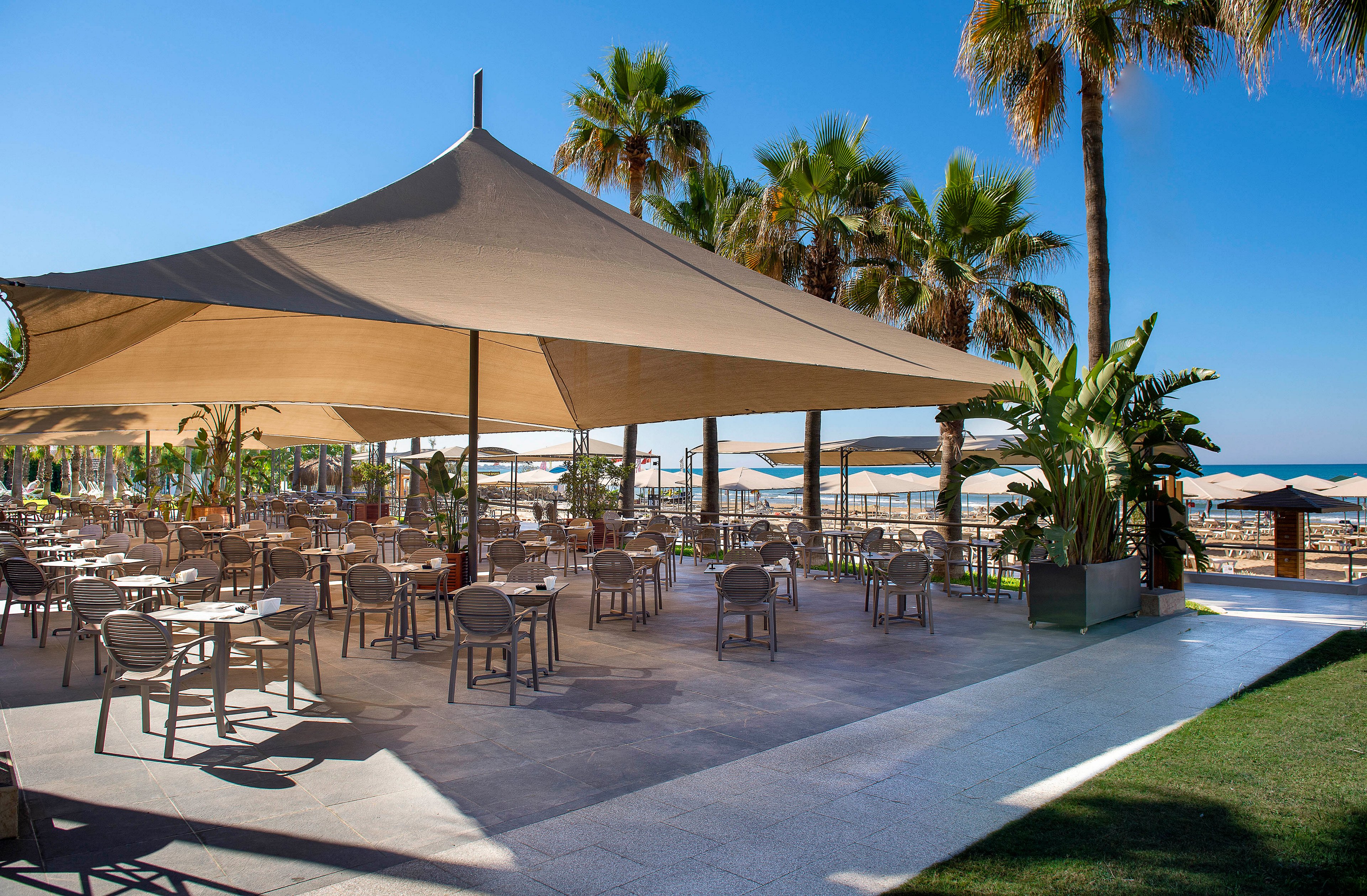 An open-air café terrace sits beside a sandy beach under a large triangular shade canopy. Numerous empty tables and chairs are arranged neatly on a tiled patio, ready for guests. Tall palm trees and lush tropical plants frame the seating area, with rows of beach umbrellas visible in the background. The clear blue sky and bright sunlight create a relaxed, vacation atmosphere by the sea.