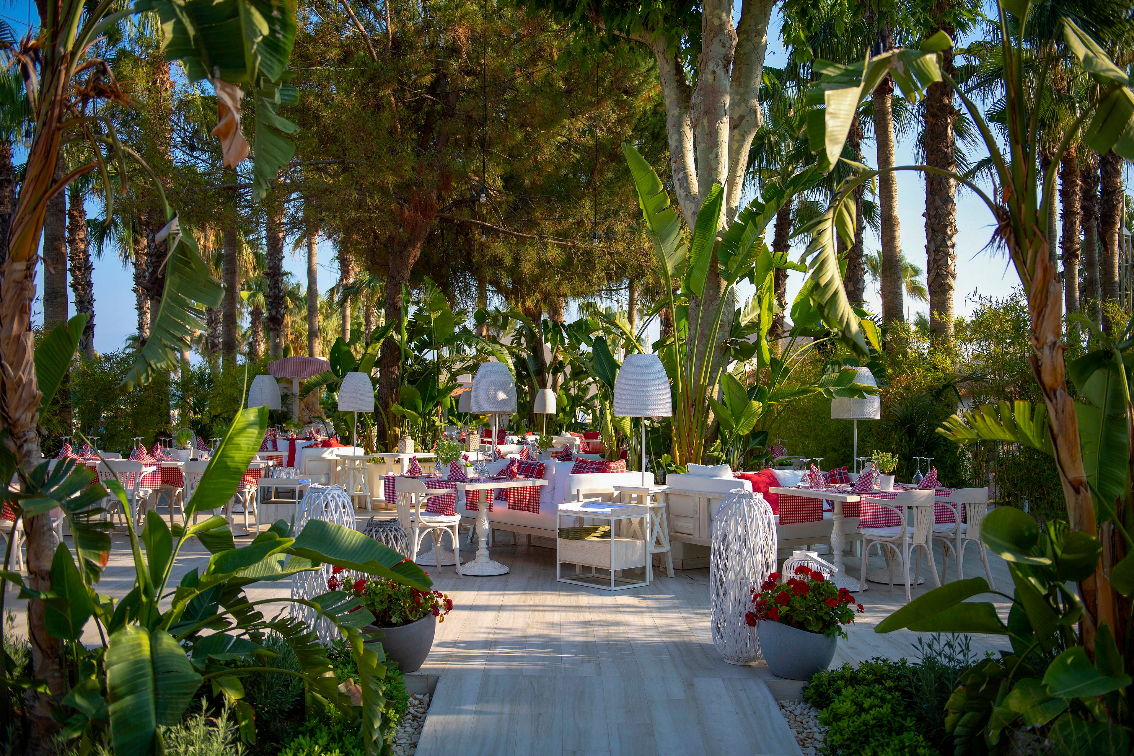 An elegant outdoor restaurant is set among lush tropical plants and tall palm trees. White tables and chairs with pink tablecloths create a vibrant and inviting dining area. Large modern floor lamps and potted flowers line the central walkway leading through the space. The scene suggests a relaxed, upscale atmosphere ideal for al fresco dining.