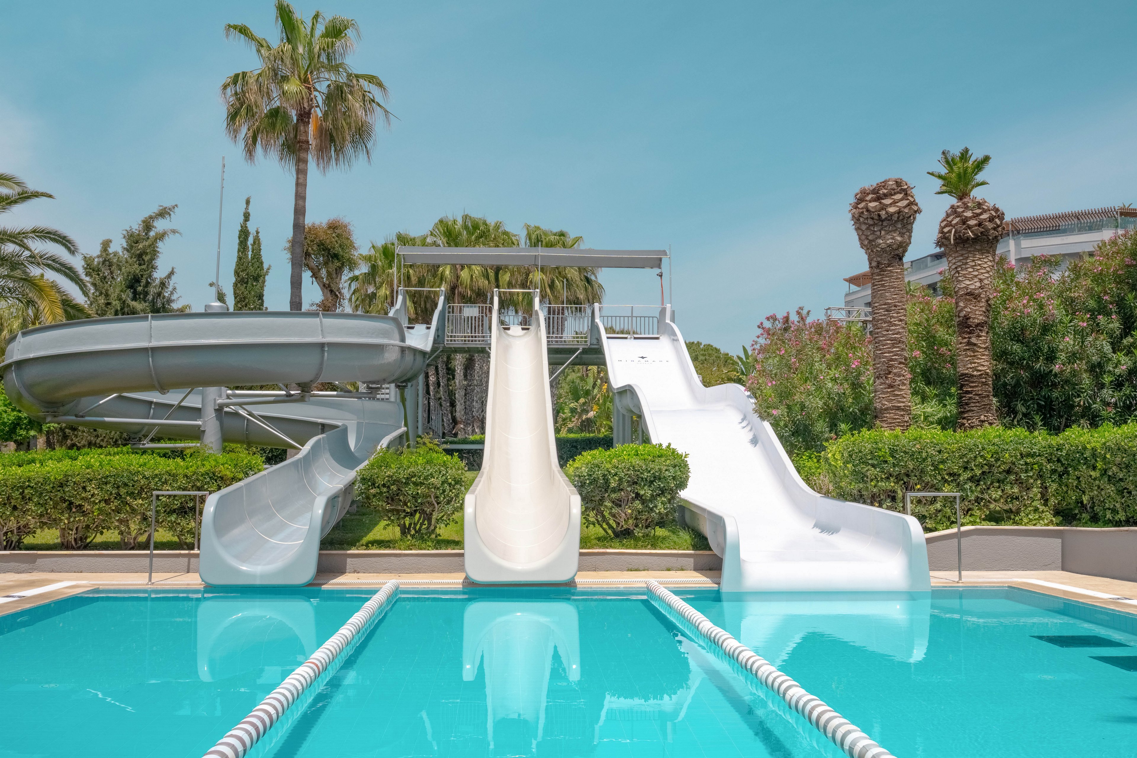 A set of three water slides leads into a bright blue swimming pool at an outdoor water park. The scene is calm and empty, with no people using the slides. Lush green hedges, palm trees, and other tropical plants surround the pool area. Clear skies and modern resort buildings in the background suggest a warm, sunny vacation setting.