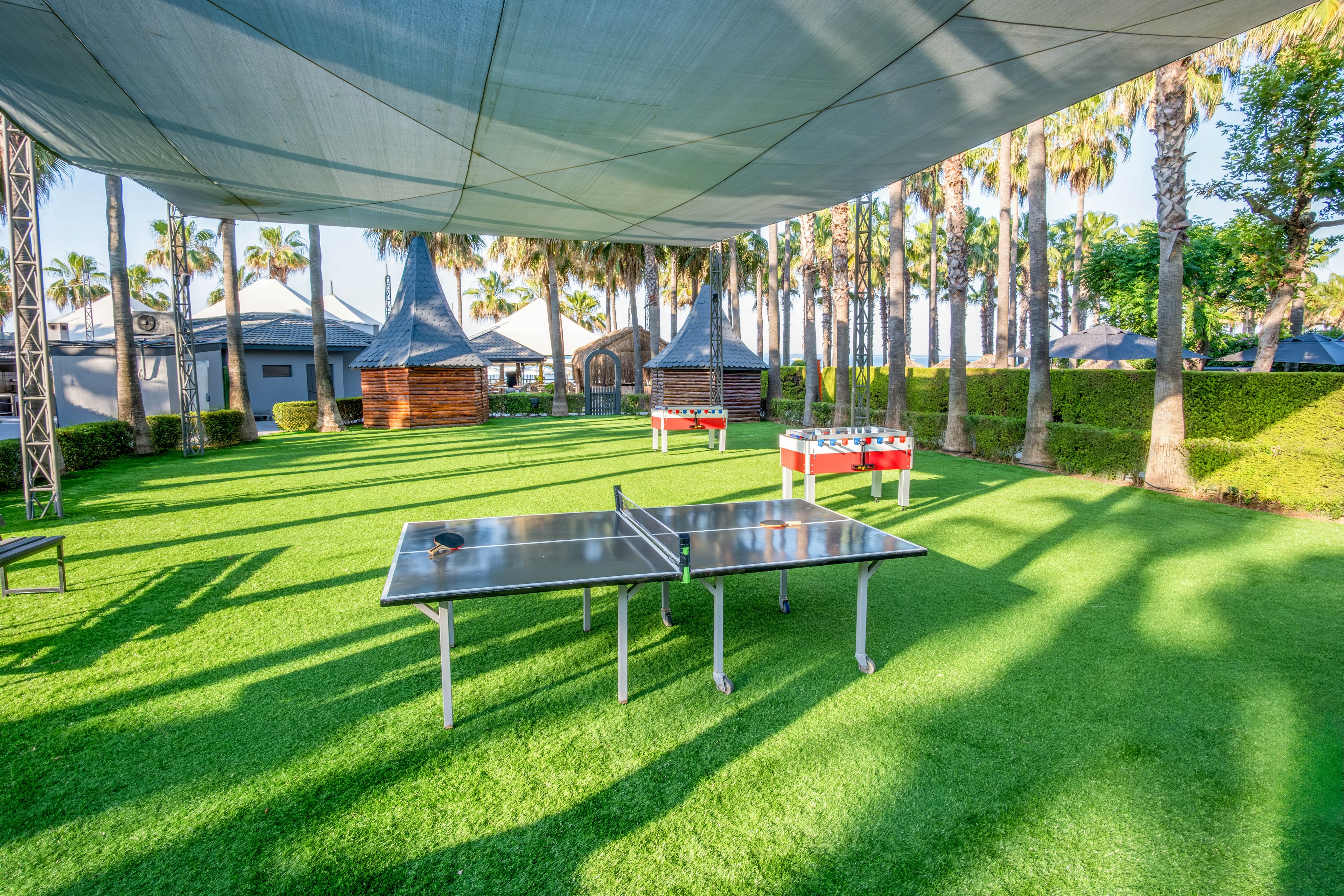 An outdoor recreation area features a table tennis table set on bright green artificial grass. In the background, a foosball table and small wooden structures sit among tall palm trees. A large canopy overhead provides shade across the play space. The scene suggests a relaxed leisure environment in a resort or park setting.