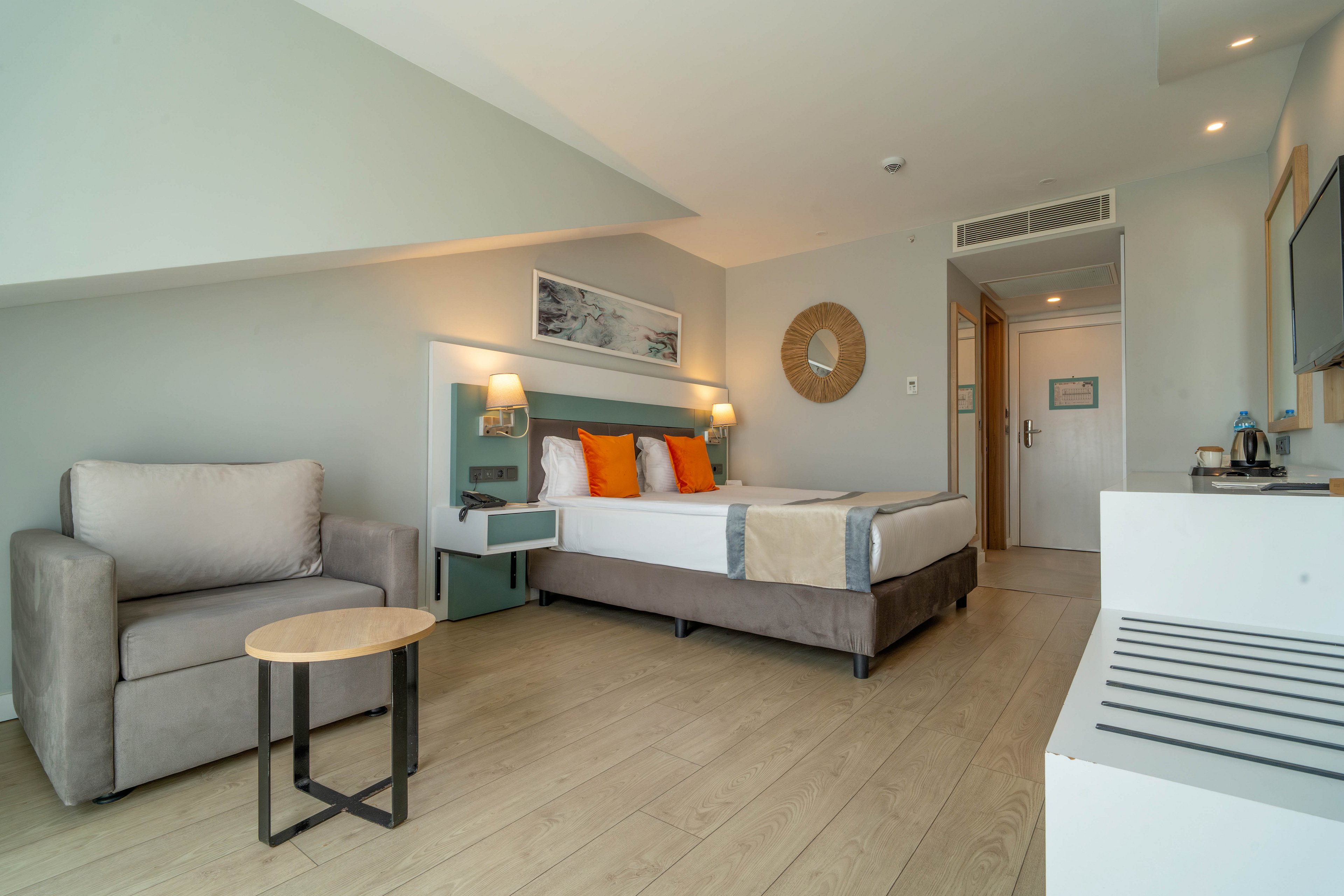 A contemporary hotel room features a double bed with white linens and orange accent pillows. The space includes a gray armchair with a small round side table and a wall-mounted television opposite the bed. Light wood flooring, soft neutral walls, and minimal artwork create a calm, airy atmosphere. A hallway with a wooden door and built-in air vent leads to the entrance and bathroom area.