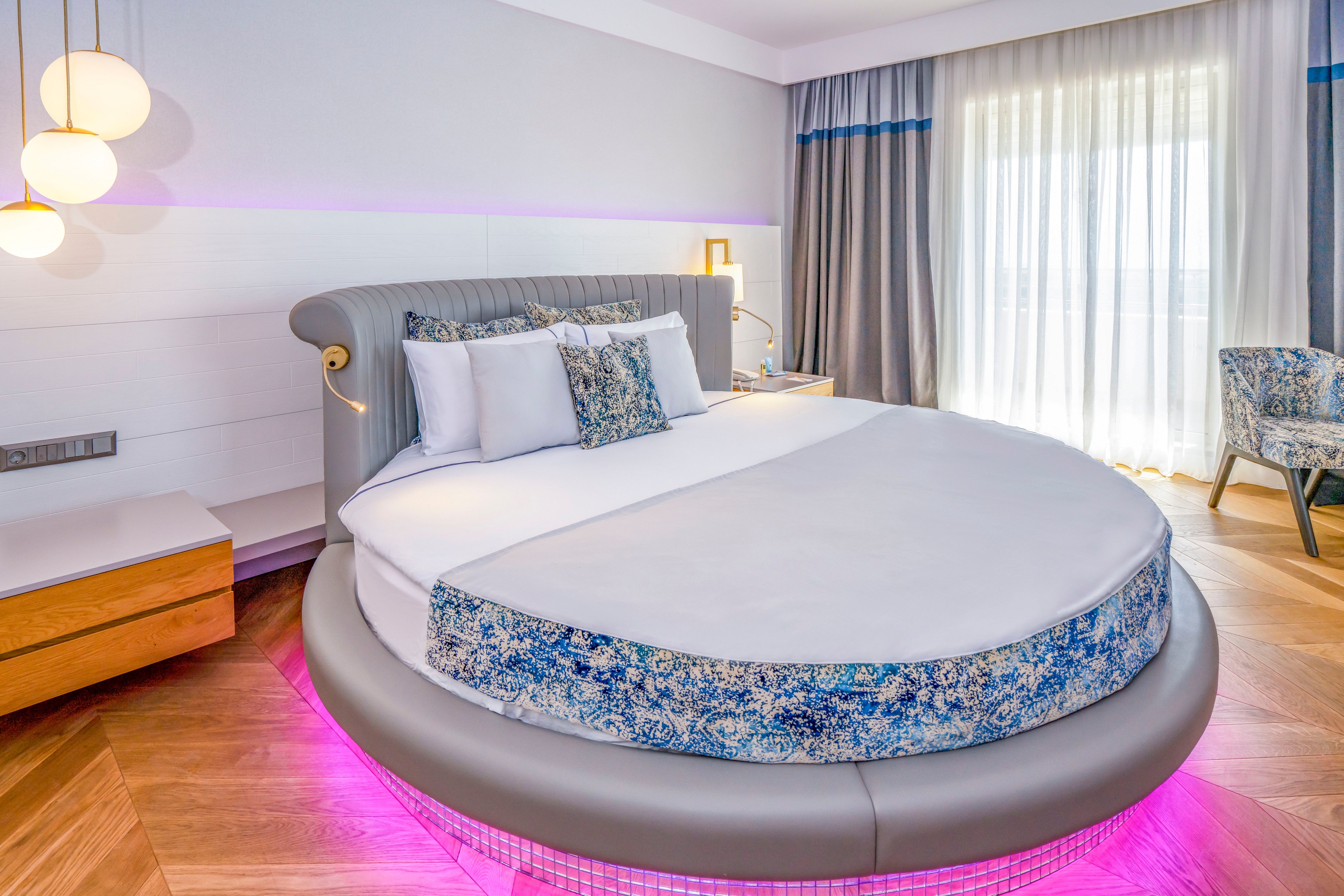 A contemporary hotel bedroom features a large round bed with white linens and patterned blue accents. Soft pink LED lighting glows beneath the bed, adding a stylish and futuristic touch to the space. Floor-to-ceiling curtains cover a bright window, complemented by a matching upholstered chair and wooden nightstand. Pendant lights and a sleek wall lamp provide warm ambient lighting, enhancing the clean and modern decor.