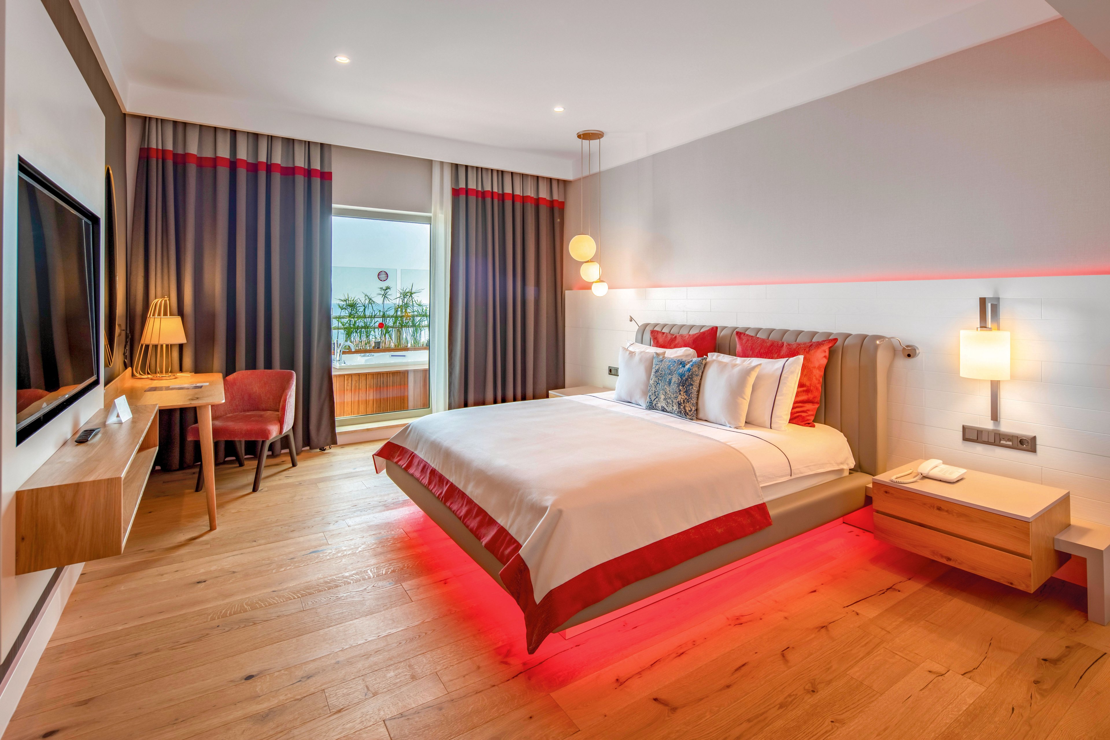 Contemporary hotel room featuring a neatly made double bed with red accent lighting beneath the frame. The space includes a wall-mounted television, bedside tables, and a small desk with a chair near the window. Floor-to-ceiling curtains frame a balcony view with outdoor seating and greenery. Warm, layered lighting and wood flooring create a cozy yet modern atmosphere.
