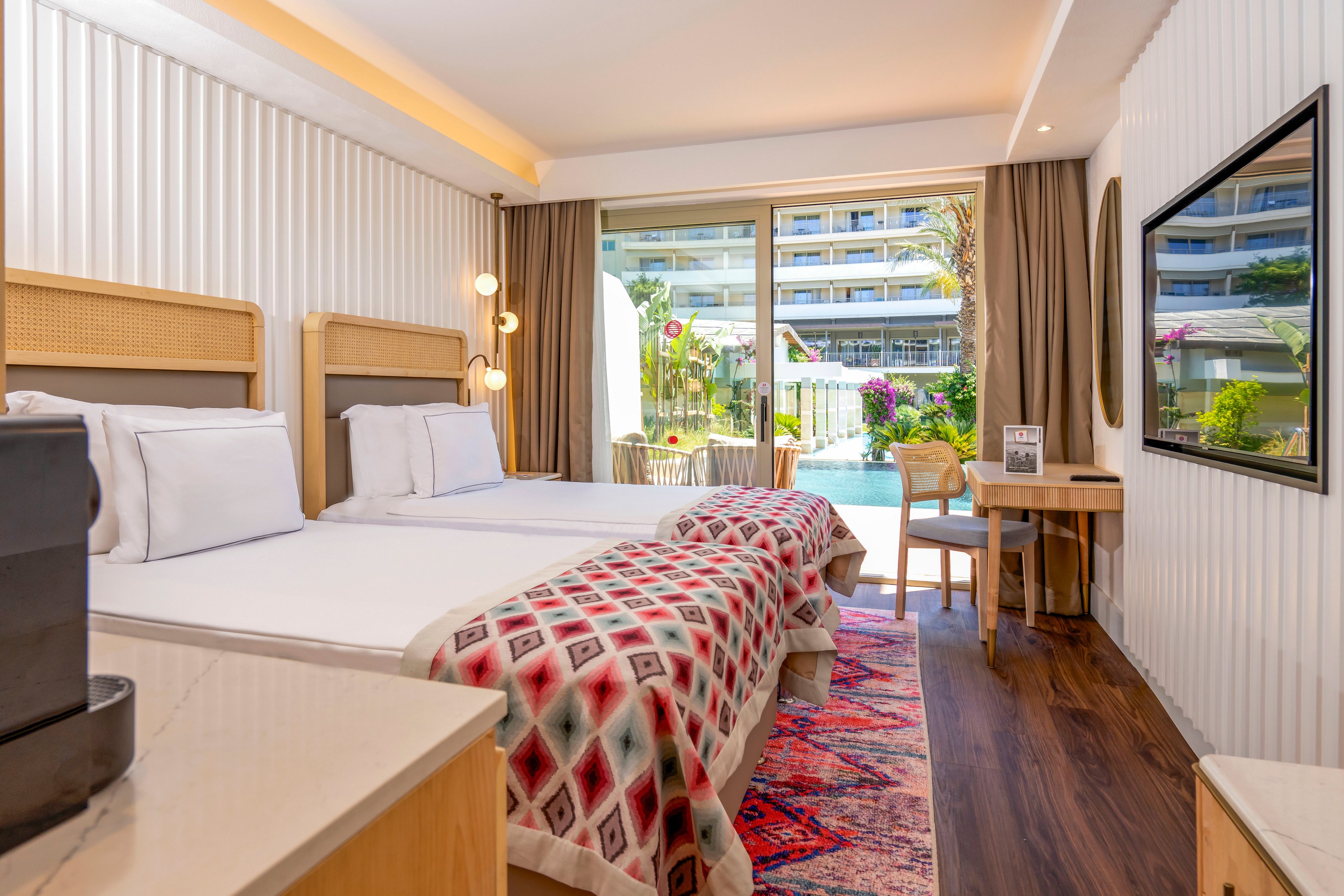 A modern hotel room features two neatly made twin beds with patterned throws and white pillows. Warm wood accents, a colorful rug, and contemporary lighting create a welcoming atmosphere. Large glass doors open to a terrace with outdoor seating, overlooking a pool and surrounding resort buildings. A desk area, television, and coffee station complete the comfortable vacation setting.