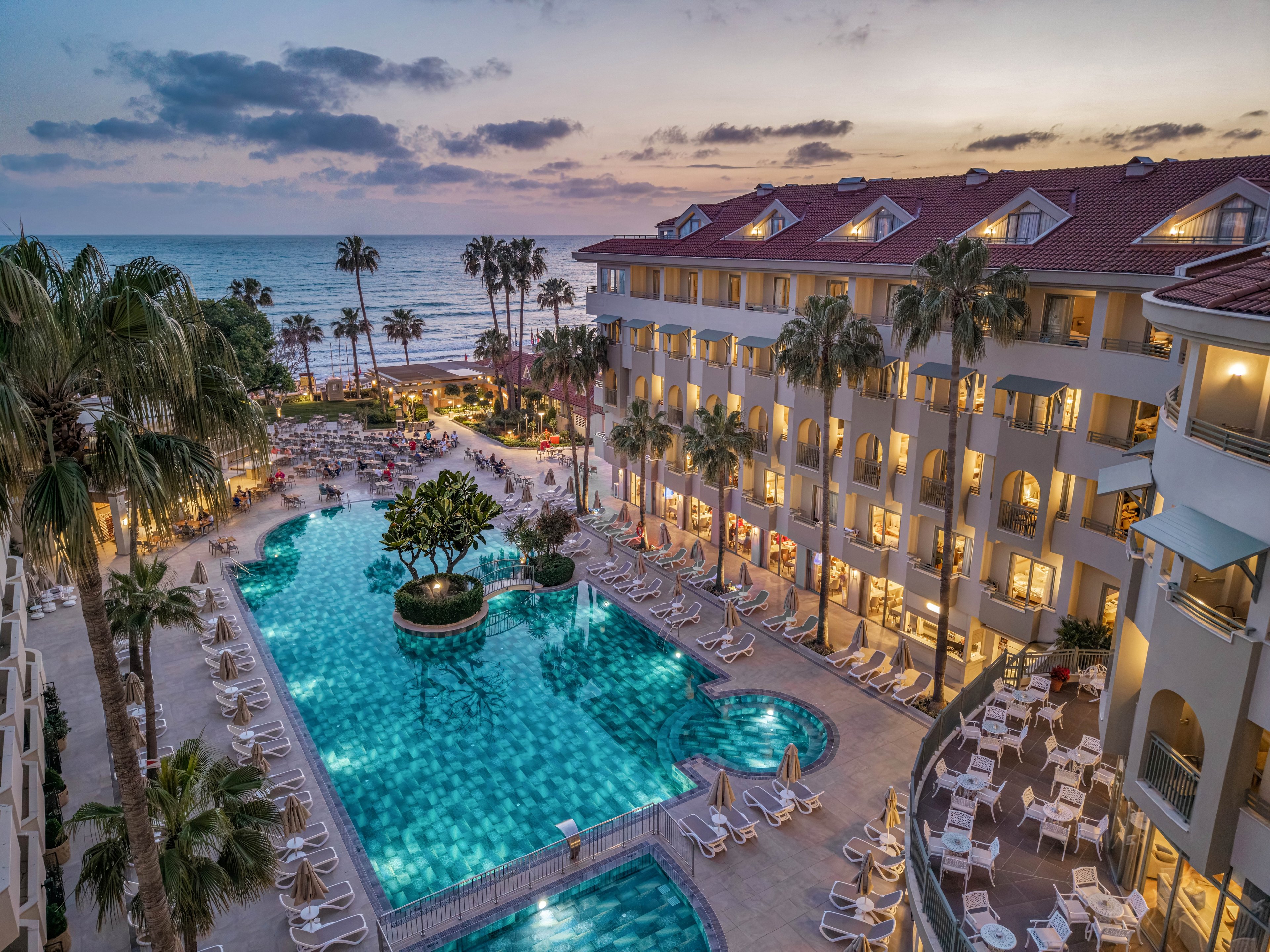 A large beachfront resort features a central swimming pool surrounded by palm trees and multi-story hotel buildings. Numerous lounge chairs and umbrellas line the pool deck, creating a relaxing vacation atmosphere. The ocean and sandy beach are visible in the background under a colorful evening sky. Soft lighting from the buildings and pool enhances the tranquil, tropical setting.