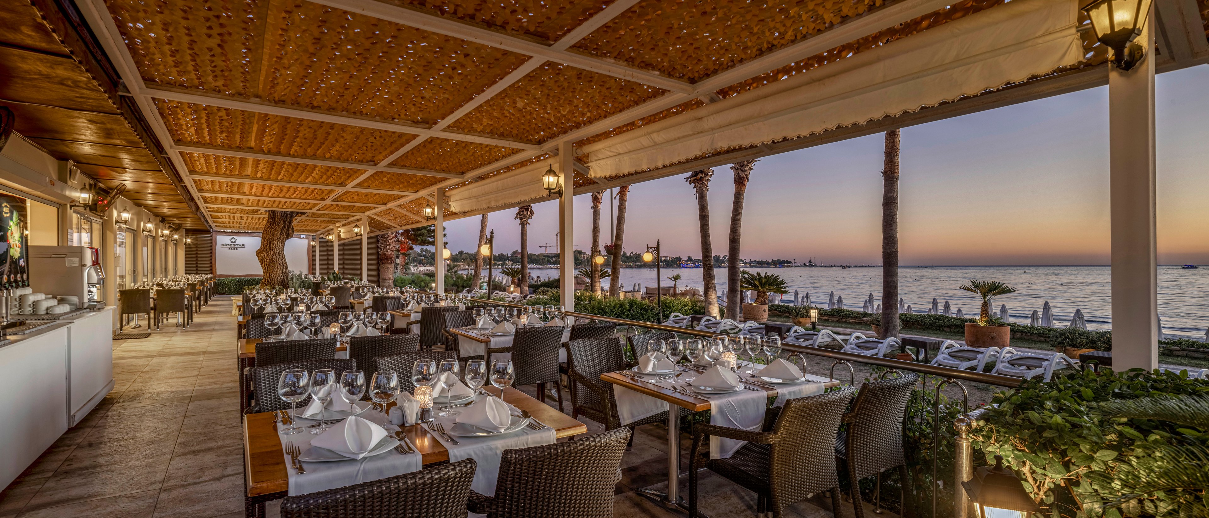 An elegant open-air restaurant terrace overlooks a calm seaside at sunset. Neatly arranged tables are set with white tablecloths, folded napkins, and polished glassware, creating a refined dining atmosphere. Warm lantern lighting and a wooden pergola ceiling add a cozy, inviting feel to the space. In the background, palm trees and lounge chairs line the shoreline as the sky glows with soft evening colors.