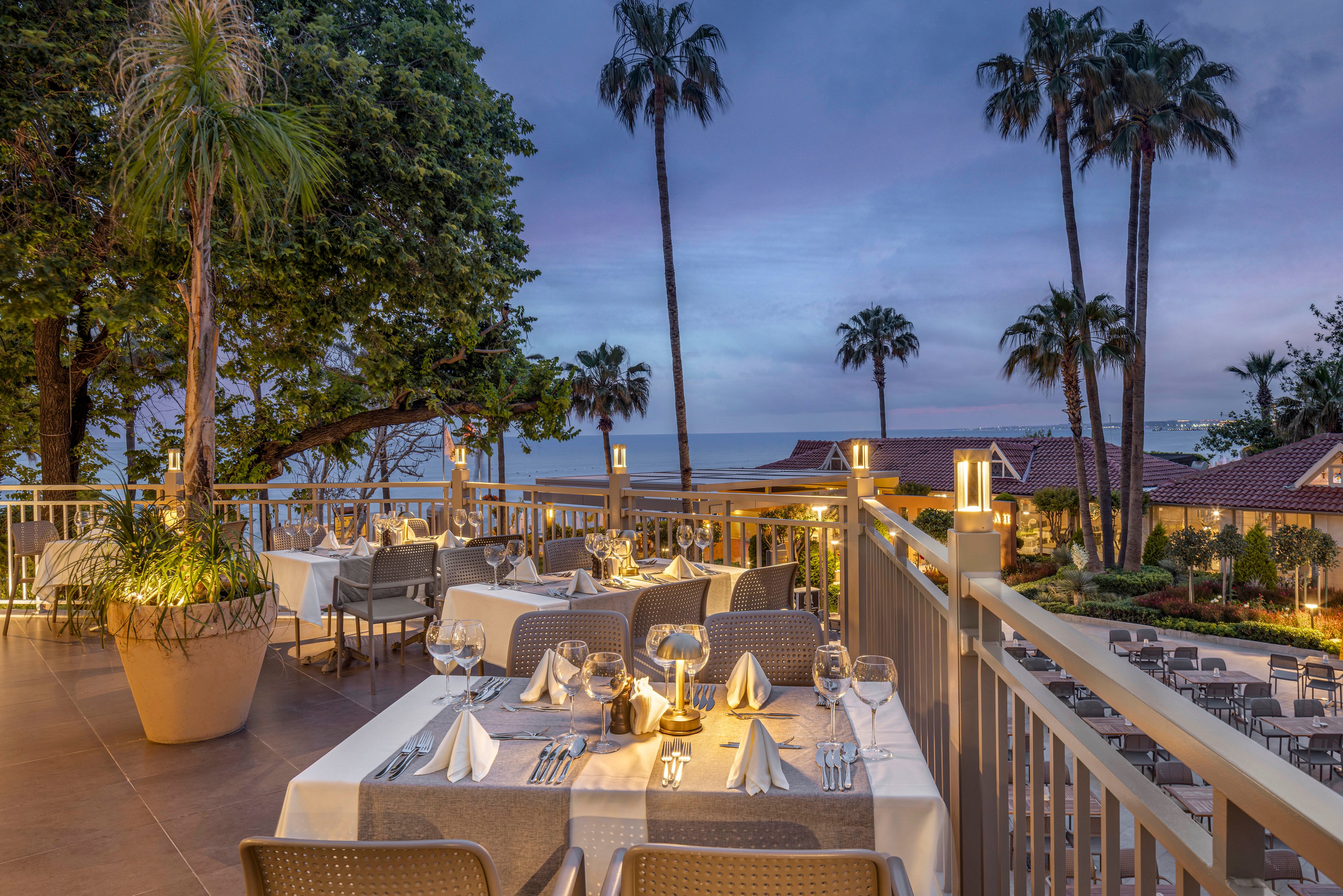 An upscale outdoor restaurant terrace is set for dinner overlooking the ocean at dusk. Tables are neatly arranged with white tablecloths, folded napkins, glassware, and candlelit centerpieces. Warm ambient lighting from lanterns and nearby buildings creates a cozy atmosphere. Tall palm trees, lush greenery, and a view of the coastline enhance the relaxed, coastal resort setting.