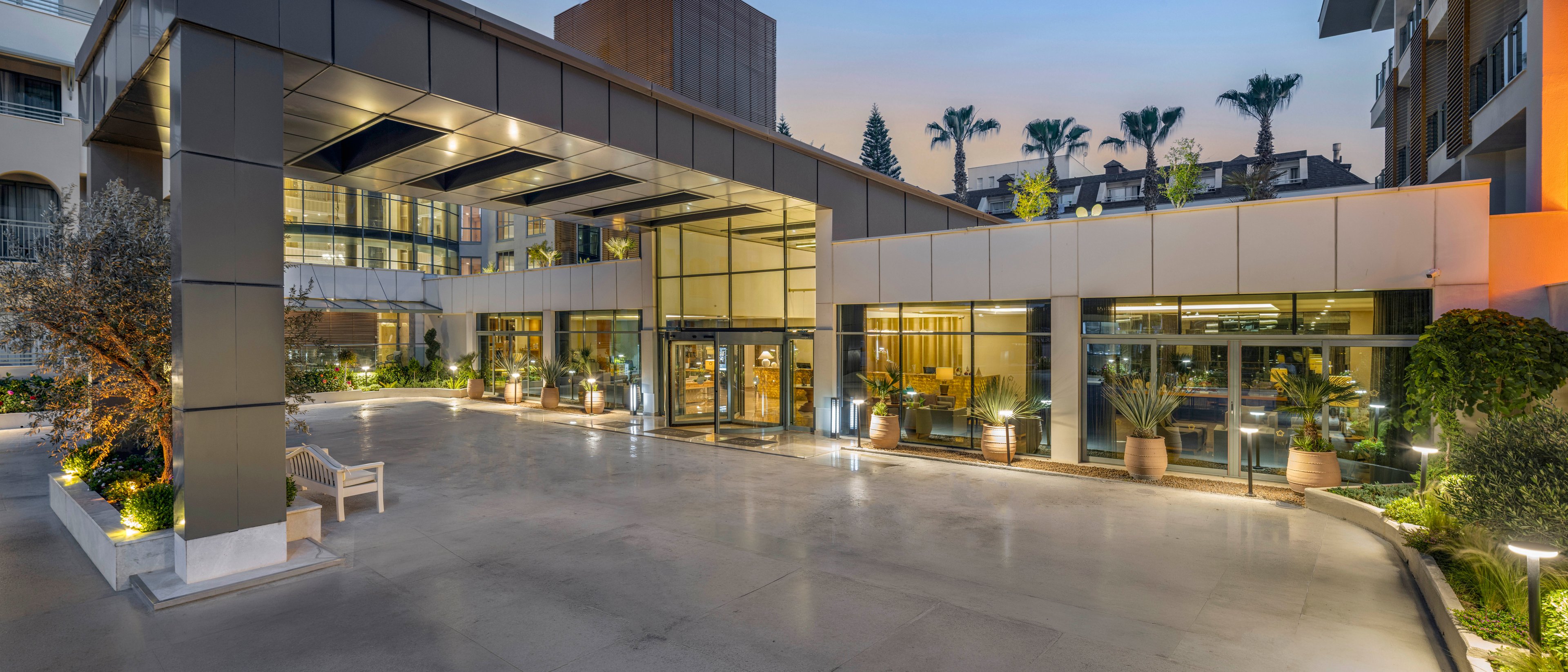 A contemporary hotel entrance is shown at dusk with a wide driveway leading to a covered drop-off area. Large glass doors and windows reveal the warmly lit interior. Decorative plants, trees, and accent lighting line the building, creating an inviting atmosphere. The sky is softly lit, and surrounding buildings and palm trees are visible in the background.