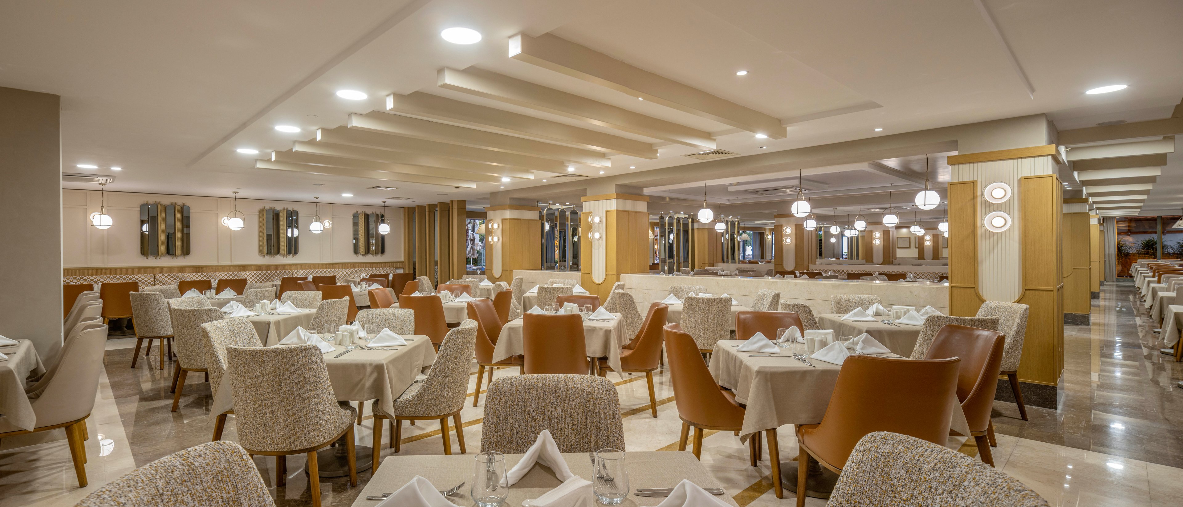 A spacious restaurant dining room is arranged with neatly set tables and upholstered chairs. White tablecloths and folded napkins create a formal, refined atmosphere. Warm neutral tones, soft ceiling lighting, and reflective wall panels contribute to a contemporary design. The room appears empty of guests, emphasizing the clean, orderly layout and inviting ambiance.
