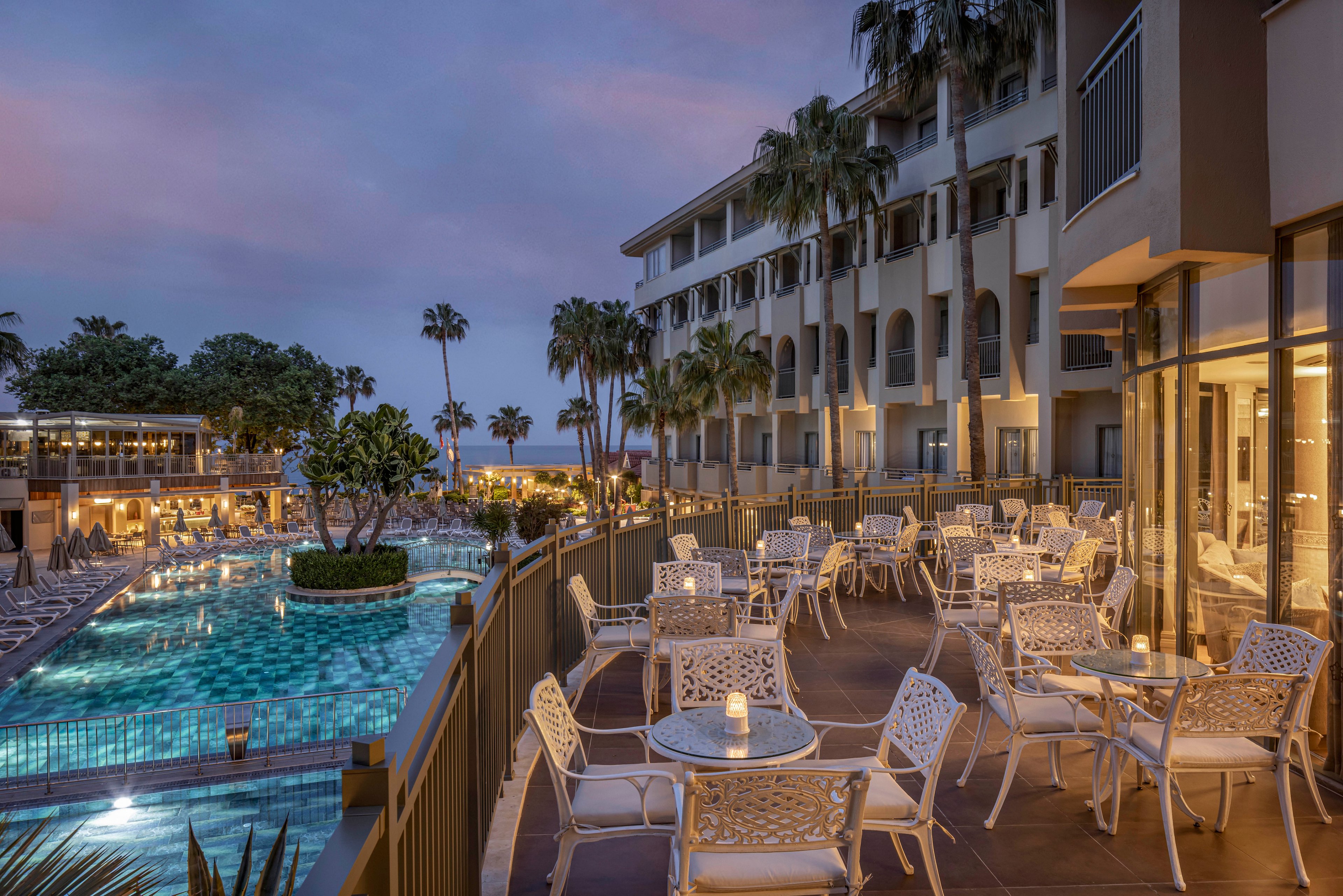 An elegant hotel terrace overlooks a large illuminated swimming pool at dusk. White metal tables and chairs are neatly arranged for outdoor dining, with small candles glowing on each table. The multi-story resort building and palm trees frame the scene under a soft purple-blue evening sky. Warm interior lights from the restaurant create a cozy, inviting atmosphere for guests.