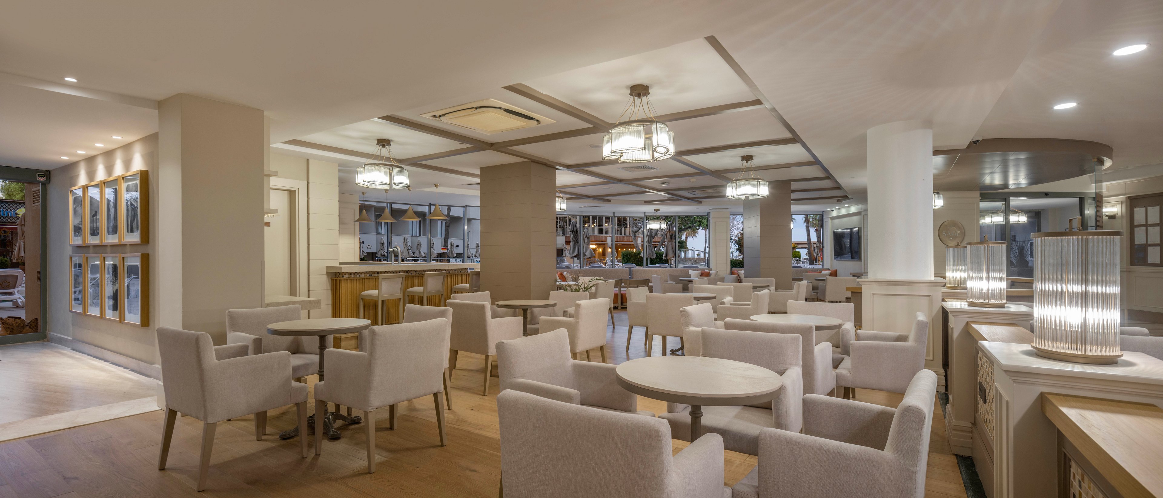 A contemporary hotel lounge features neatly arranged round tables and cushioned armchairs in soft neutral tones. The space is open and well-lit, with ceiling lights and decorative chandeliers creating a warm ambiance. A bar area is visible in the background, adding to the social and hospitality-focused setting. Large windows and reflective surfaces contribute to a spacious, airy feel.