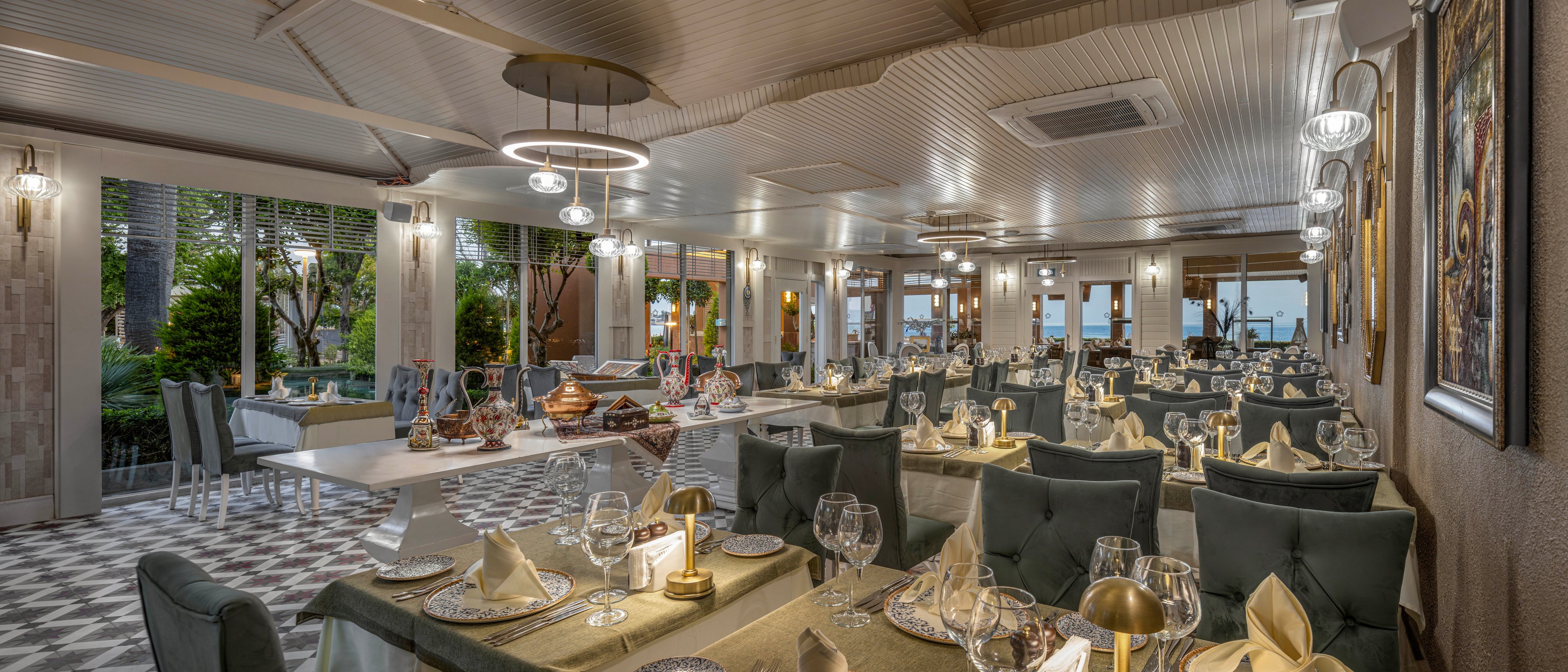 A spacious, upscale restaurant dining room is set with neatly arranged tables and folded napkins. Glassware, cutlery, and decorative centerpieces create a refined atmosphere. Large windows reveal greenery and a glimpse of the sea outside, adding a coastal feel. Warm lighting from modern chandeliers and wall sconces enhances the sophisticated, inviting ambiance.