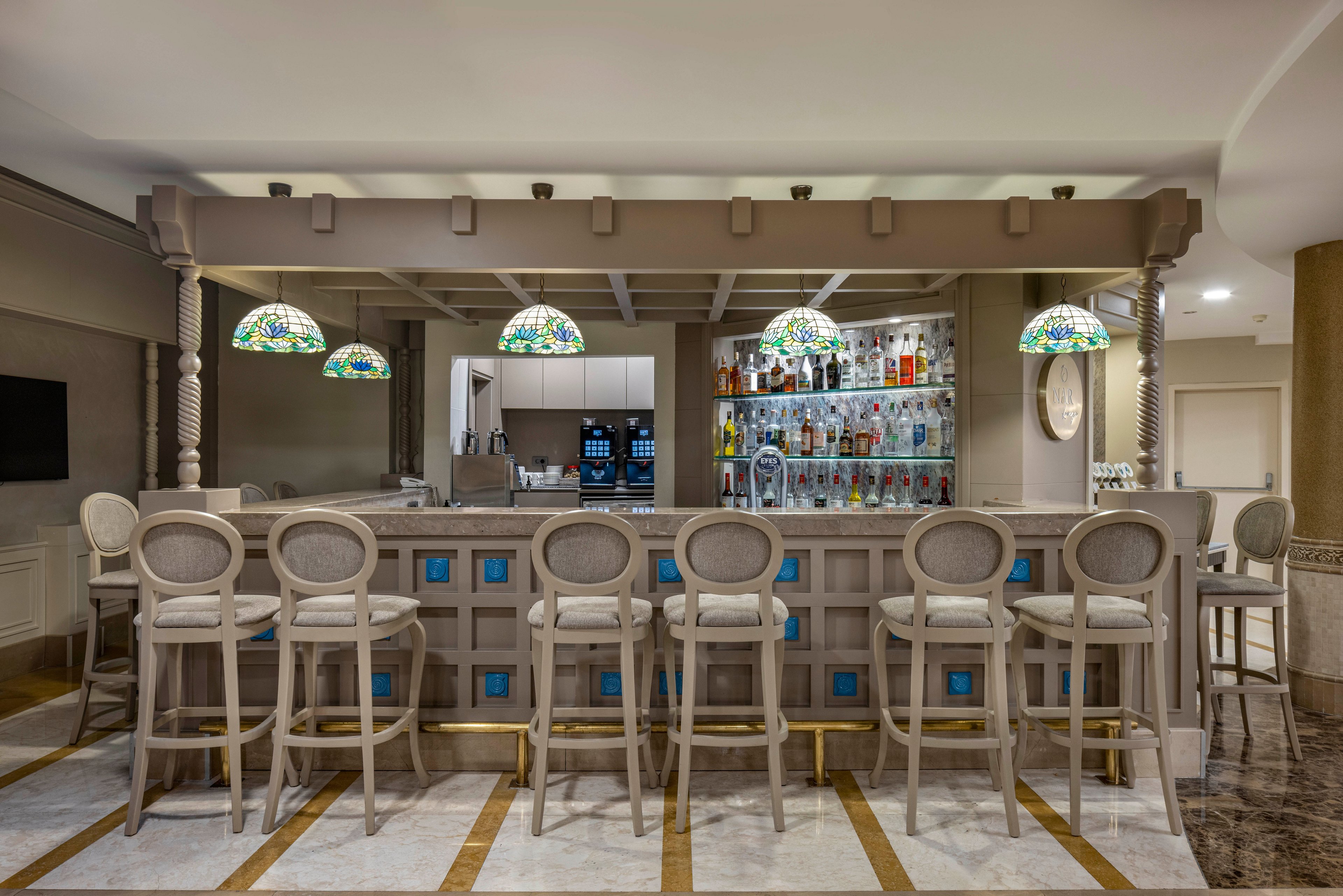 A stylish hotel bar features a row of high-backed stools facing a marble countertop. Behind the bar, shelves are filled with assorted liquor bottles and glassware, creating a colorful focal point. Decorative stained-glass pendant lamps hang above the counter, casting a warm, inviting glow. The neutral-toned interior and clean lines give the space a modern yet classic atmosphere.