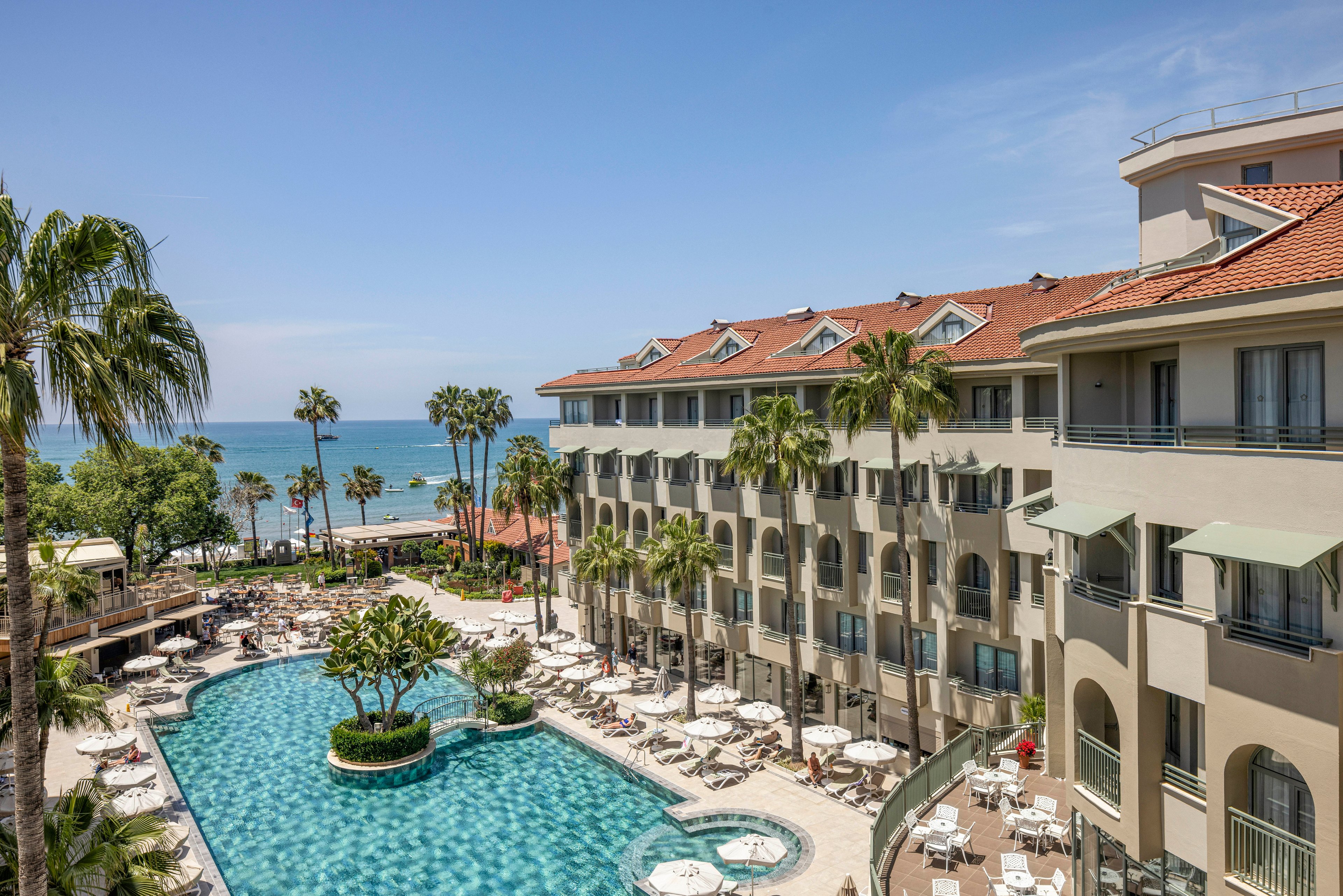 A large beachfront resort hotel features a spacious outdoor swimming pool surrounded by sun loungers and umbrellas. The multi-story building with red-tiled roofs overlooks the pool area and the nearby sea. Palm trees and landscaped greenery add a tropical atmosphere to the scene. Clear blue skies and calm ocean water suggest a warm, sunny vacation destination.