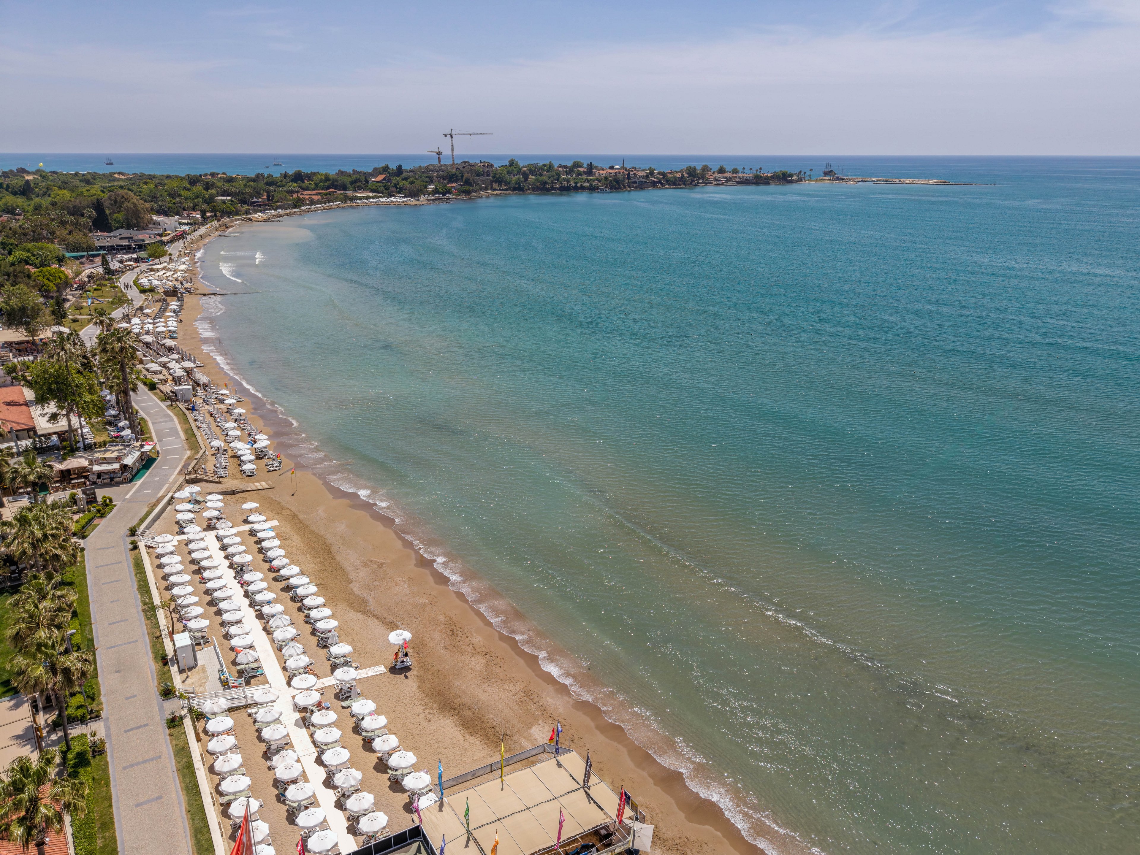 An aerial view shows a long sandy beach lined with rows of white sun umbrellas and loungers. A calm turquoise sea stretches out to the horizon, gently meeting the shoreline. A paved promenade with palm trees and buildings runs parallel to the beach. In the distance, a small peninsula and light haze complete the tranquil coastal scene.
