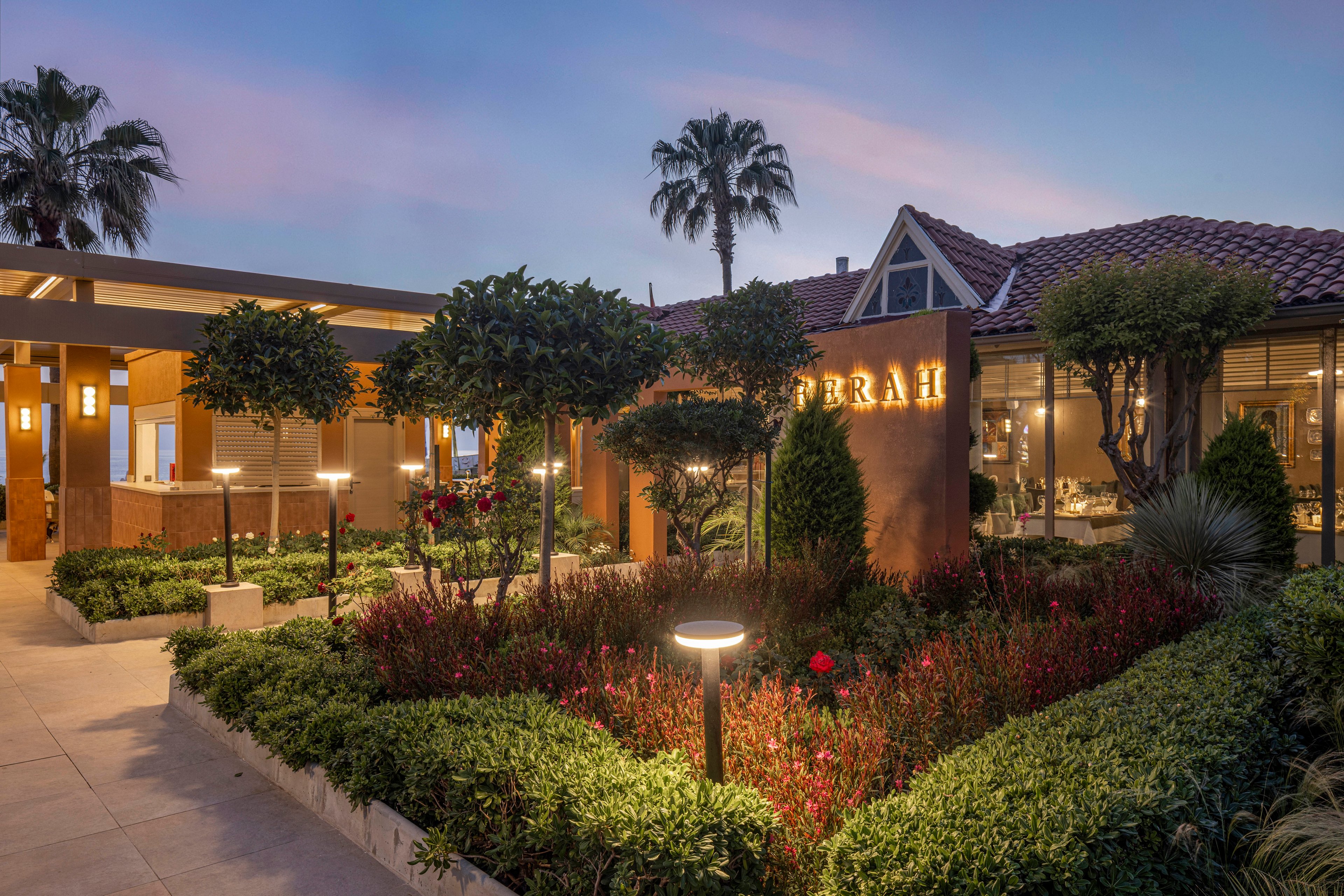 A contemporary restaurant entrance is shown at dusk with warm interior lighting visible through large windows. The foreground features a landscaped garden with manicured shrubs, flowering plants, and pathway lights. Palm trees and a tiled roof frame the building against a soft evening sky. A tall sign with illuminated text stands near the walkway, enhancing the upscale and inviting atmosphere.