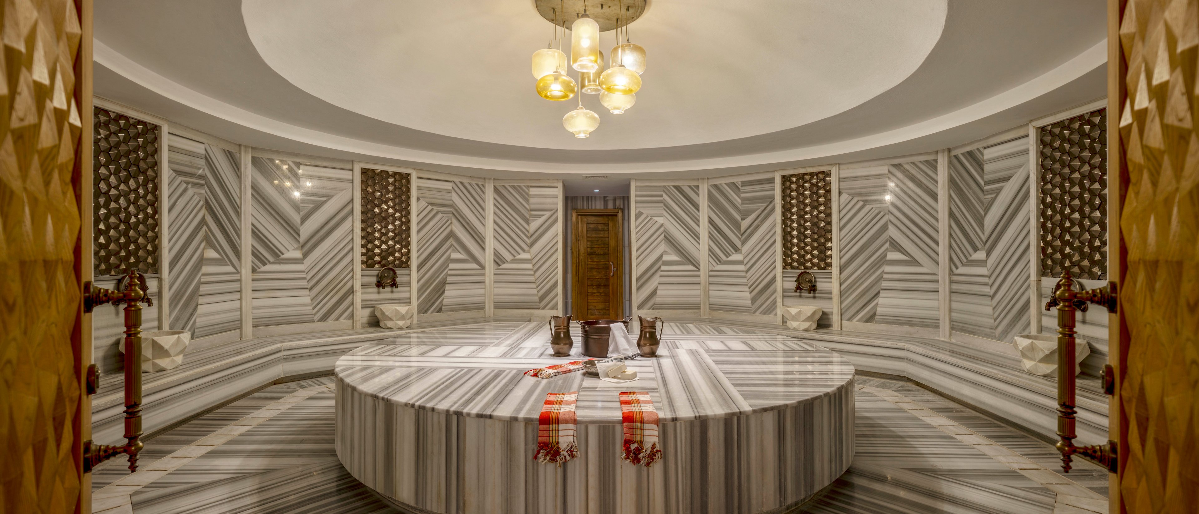 A luxurious circular Turkish hammam features a large central marble platform surrounded by continuous bench seating. The room is clad in striped marble panels arranged in geometric patterns, creating a sophisticated spa ambiance. Warm pendant lights hang from the ceiling, casting a soft glow over the stone surfaces. Traditional towels and cushions are placed on the central slab, emphasizing relaxation and wellness in this serene bath setting.