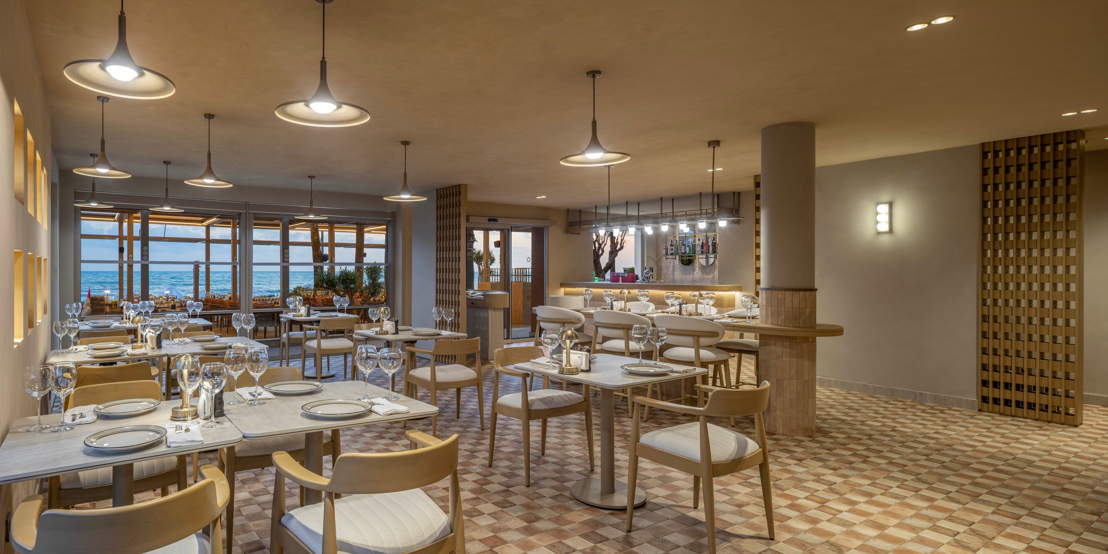 A contemporary restaurant dining room features neatly arranged tables set with plates, glasses, and cutlery. Light wooden chairs and neutral-toned furniture create a calm, minimalist atmosphere. Pendant lights hang from the ceiling, casting a warm glow over the patterned tile floor. Large windows at the back reveal a view toward the sea and the fading daylight outside.