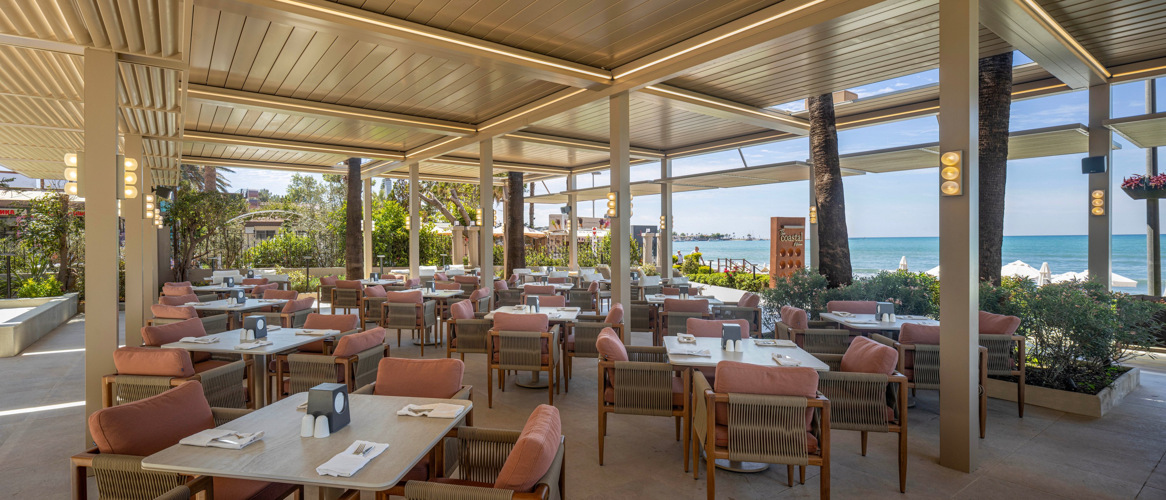 An open-air restaurant with neatly arranged tables and cushioned chairs overlooks a bright blue ocean. The covered terrace features a modern pergola-style roof that shades diners from the sun. Palm trees and lush greenery frame the seating area, adding a tropical resort atmosphere. Soft neutral tones and pink upholstery create a relaxed, coastal dining ambiance.