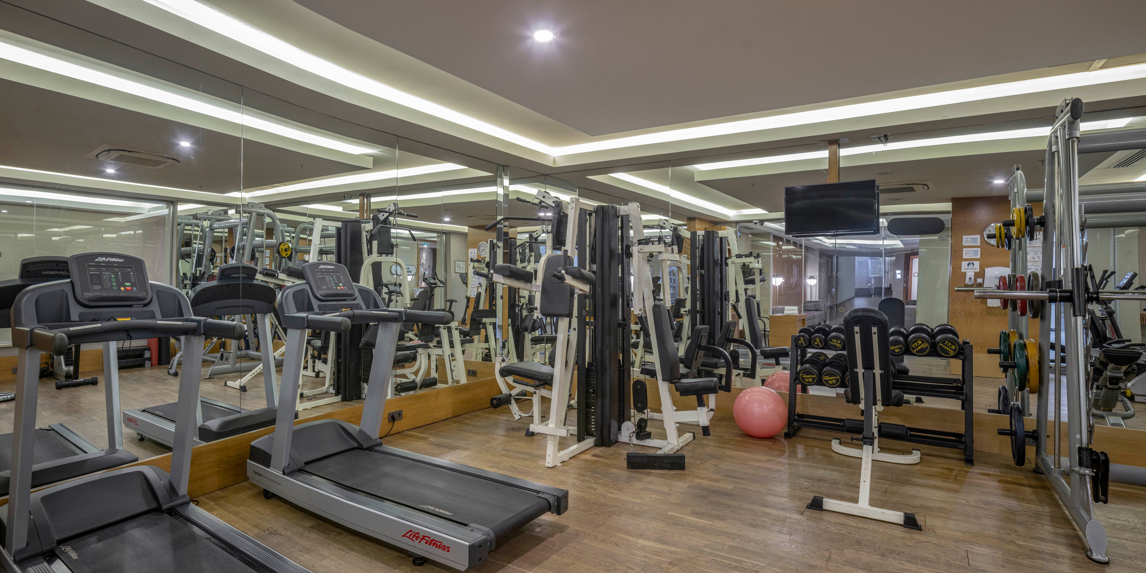 A modern indoor fitness center features multiple treadmills, weight machines, and free weights neatly arranged on a wooden floor. Large wall mirrors expand the sense of space and reflect the equipment throughout the room. Overhead recessed lighting and a mounted television create a bright, contemporary workout environment. A pink exercise ball and organized dumbbell racks add variety to the available training options.
