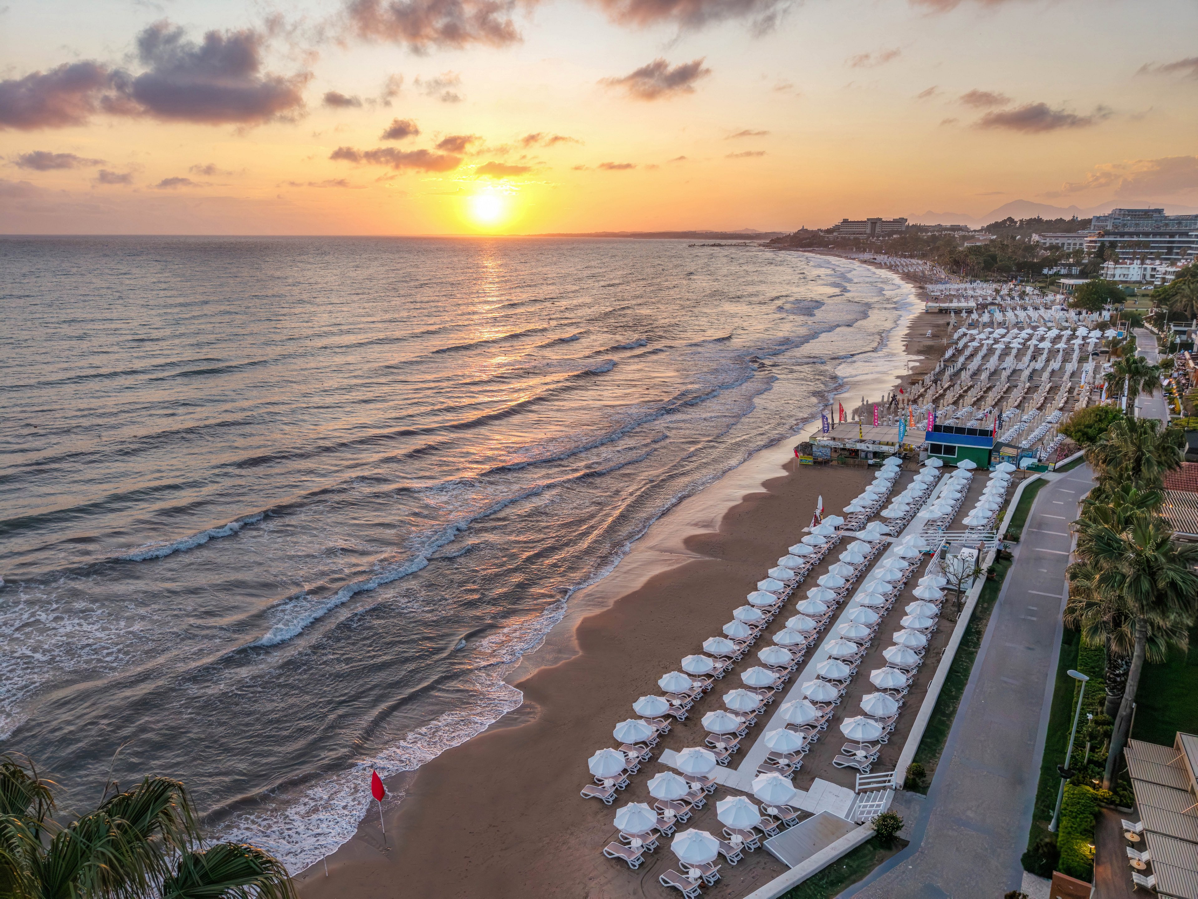 A tranquil sunset illuminates a long sandy beach lined with rows of white sun loungers and umbrellas. Gentle waves roll onto the shore as the sky glows with warm orange and pink tones. A seaside resort with palm trees and buildings stretches along the coastline in the background. The scene conveys a peaceful vacation atmosphere at the water’s edge.