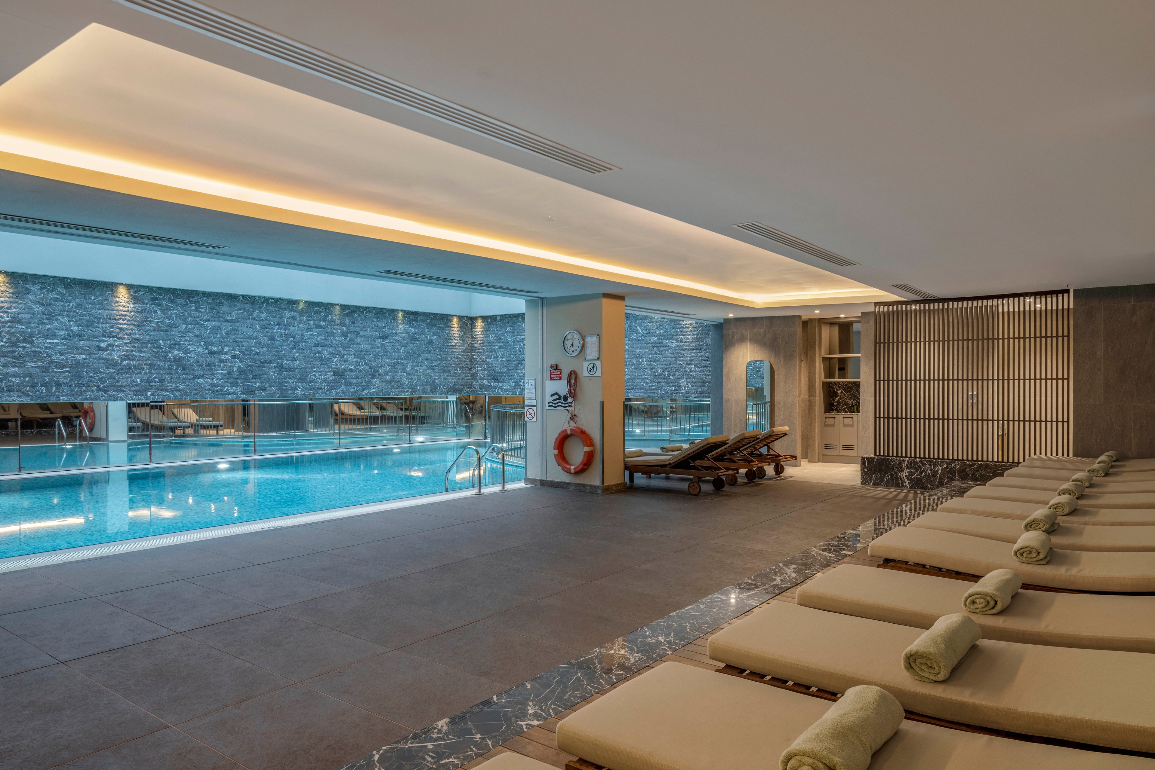 A contemporary indoor spa area features a long swimming pool beside a row of cushioned loungers. Rolled towels are neatly placed on each lounger, suggesting a relaxing wellness experience. The pool is framed by stone-textured walls, soft ceiling lighting, and large windows that reflect the water. Minimalist decor and neutral tones create a calm, luxurious atmosphere.