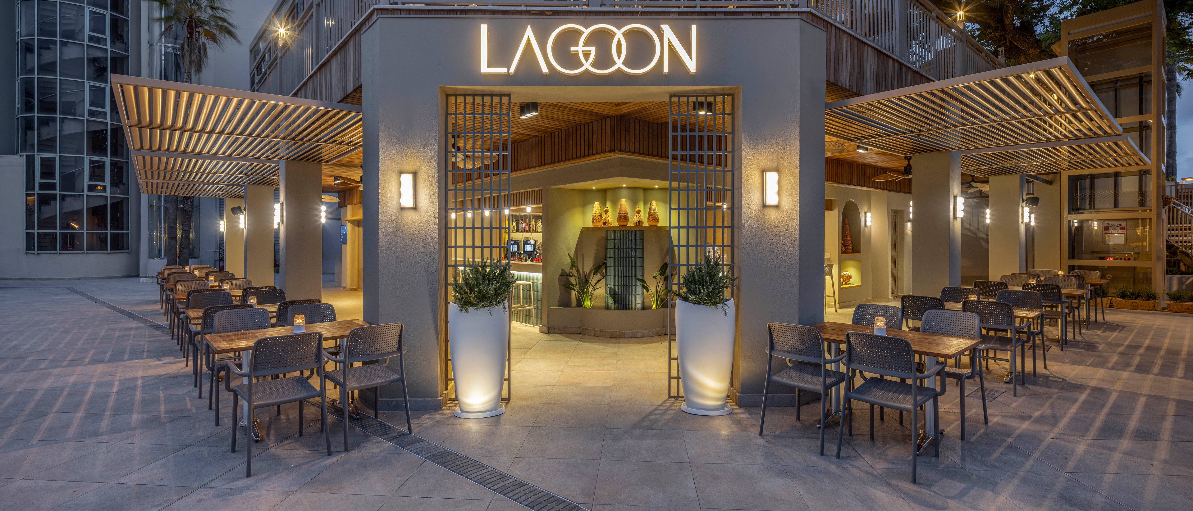 A contemporary open-air café named LAGOON is shown at dusk with warm ambient lighting. The entrance features large columns, wooden slatted pergolas, and neatly arranged outdoor seating on both sides. Inside, a bar area with illuminated shelves and decorative bottles is visible. The scene is calm and inviting, with a clean, modern architectural style and no people present.