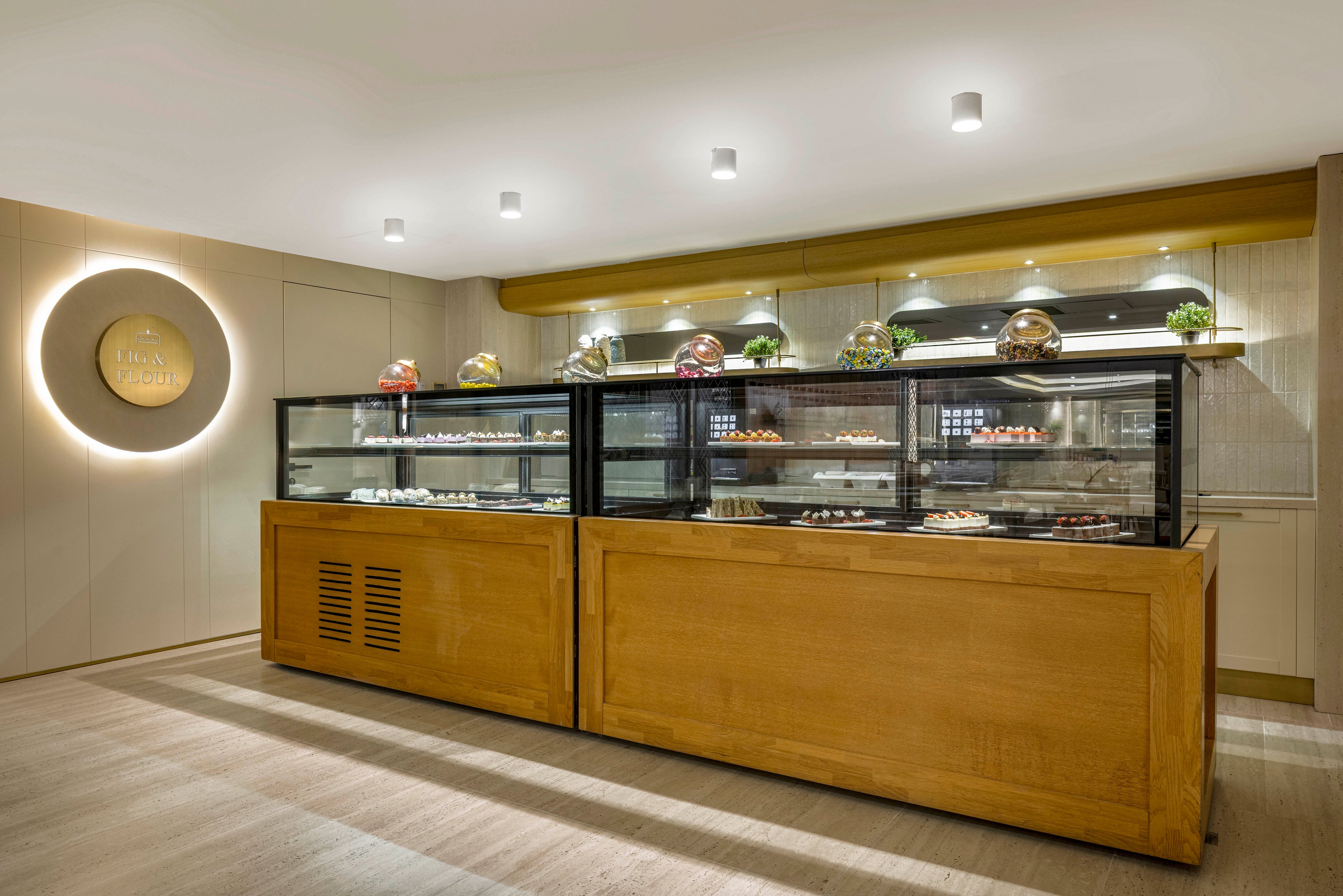 A sleek pastry display counter stands in the center of a modern bakery interior. Glass cases showcase an assortment of desserts and pastries arranged on multiple shelves. Warm wooden cabinetry contrasts with the light-colored walls and flooring. Recessed ceiling lights and under-shelf lighting highlight the treats and create a clean, inviting atmosphere. A circular wall feature with subtle text adds a decorative focal point behind the counter.