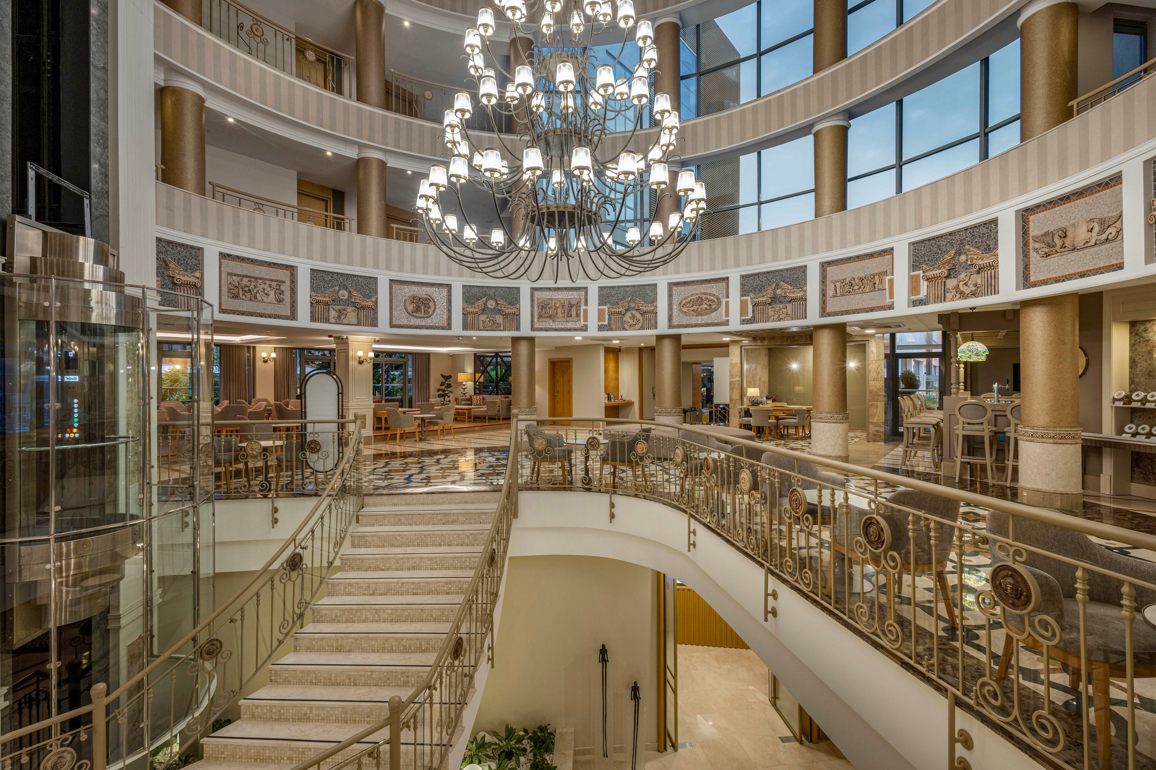 A luxurious multi-story hotel atrium features a sweeping staircase and an elaborate central chandelier. Curved balconies with decorative railings overlook the open lobby space below. Large windows allow natural light to illuminate the polished floors and elegant seating areas. Classical artwork panels and warm lighting contribute to an upscale, sophisticated atmosphere.