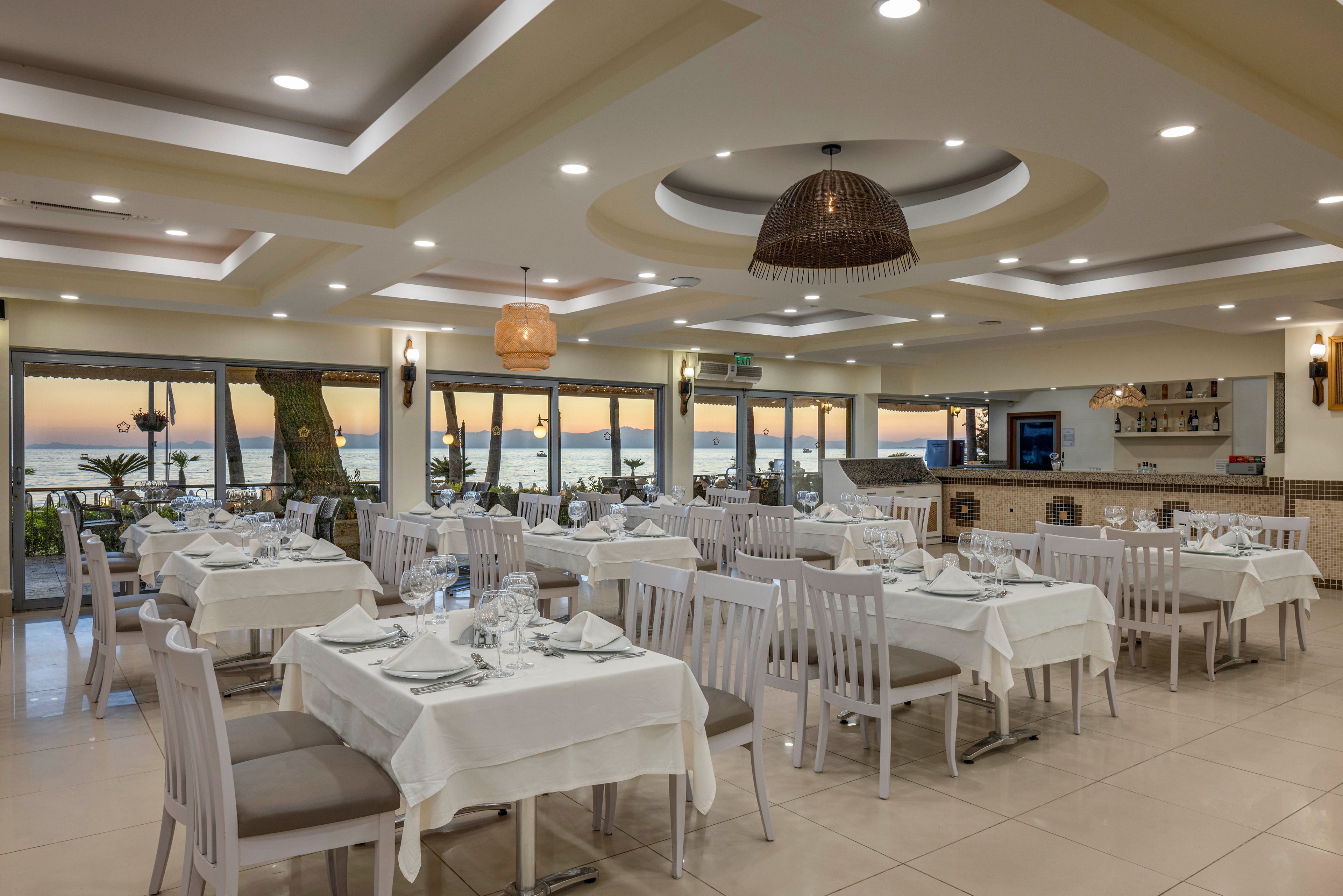 A bright, elegant restaurant dining room is set with neatly arranged white tables and chairs. Large windows reveal a scenic seaside view with palm trees and a colorful sunset over the water. Warm ceiling lights and decorative pendant lamps create a cozy, inviting atmosphere. The space appears ready for guests, with glassware and table settings carefully placed on each table.