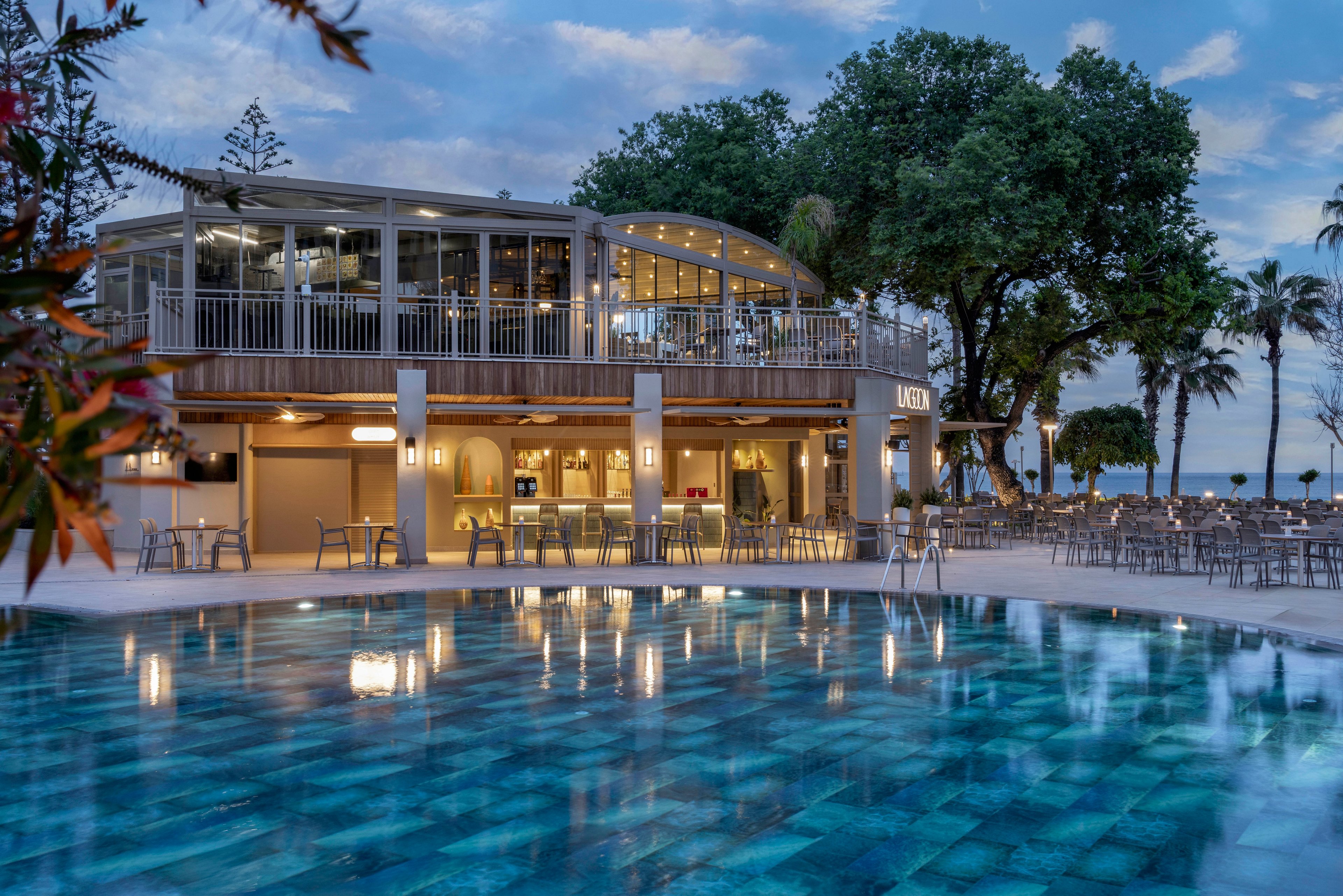 A contemporary two-story restaurant stands beside a calm, illuminated swimming pool at dusk. The building features large windows, warm interior lighting, and an upper-level terrace with railings and seating. Numerous tables and chairs are arranged around the pool and patio area, suggesting an outdoor dining or lounge space. Tall trees and glimpses of the ocean in the background create a relaxed coastal atmosphere.