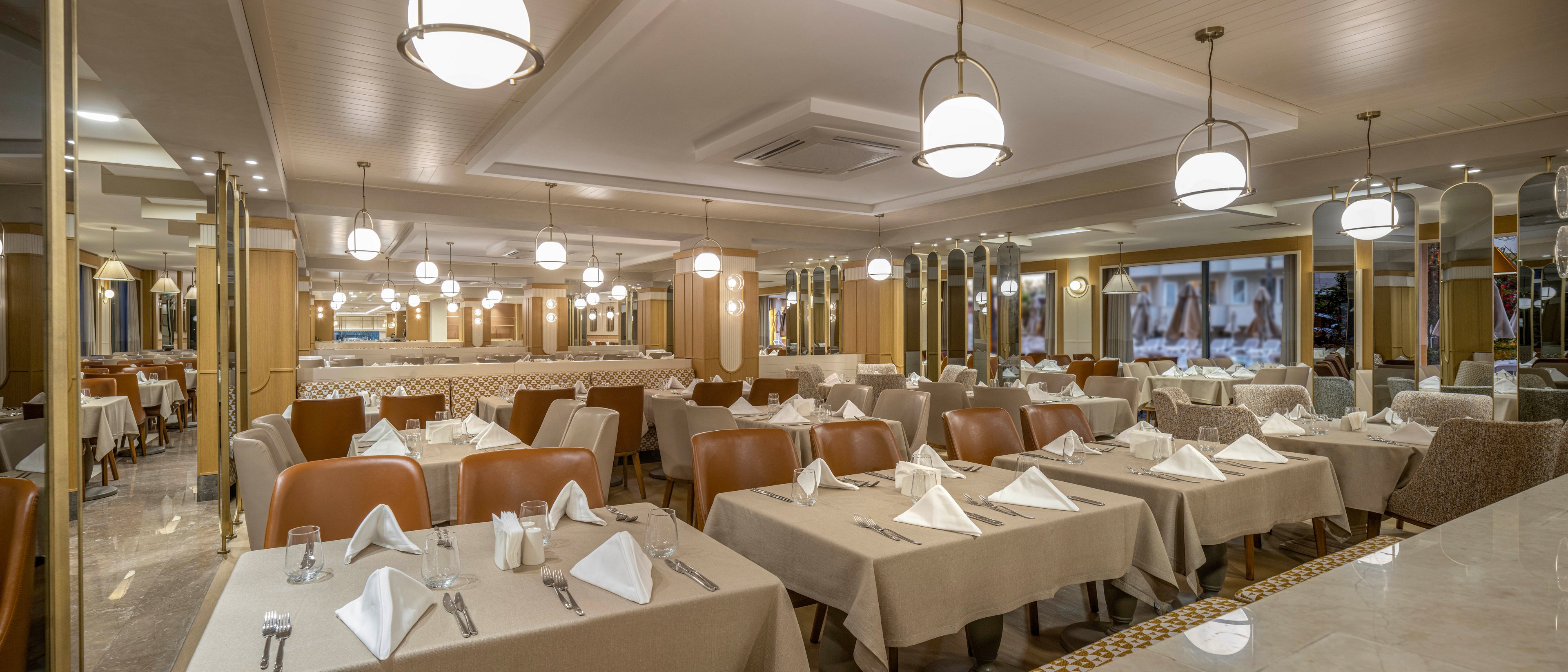 Spacious restaurant dining room with neatly arranged tables and chairs is shown empty and ready for guests. Each table is covered with a beige tablecloth and set with folded white napkins, cutlery, and small condiment shakers. Warm pendant lights and ceiling fixtures create a soft, inviting ambiance across the room. Large mirrors and wall decor enhance the sense of space and sophistication in the interior.
