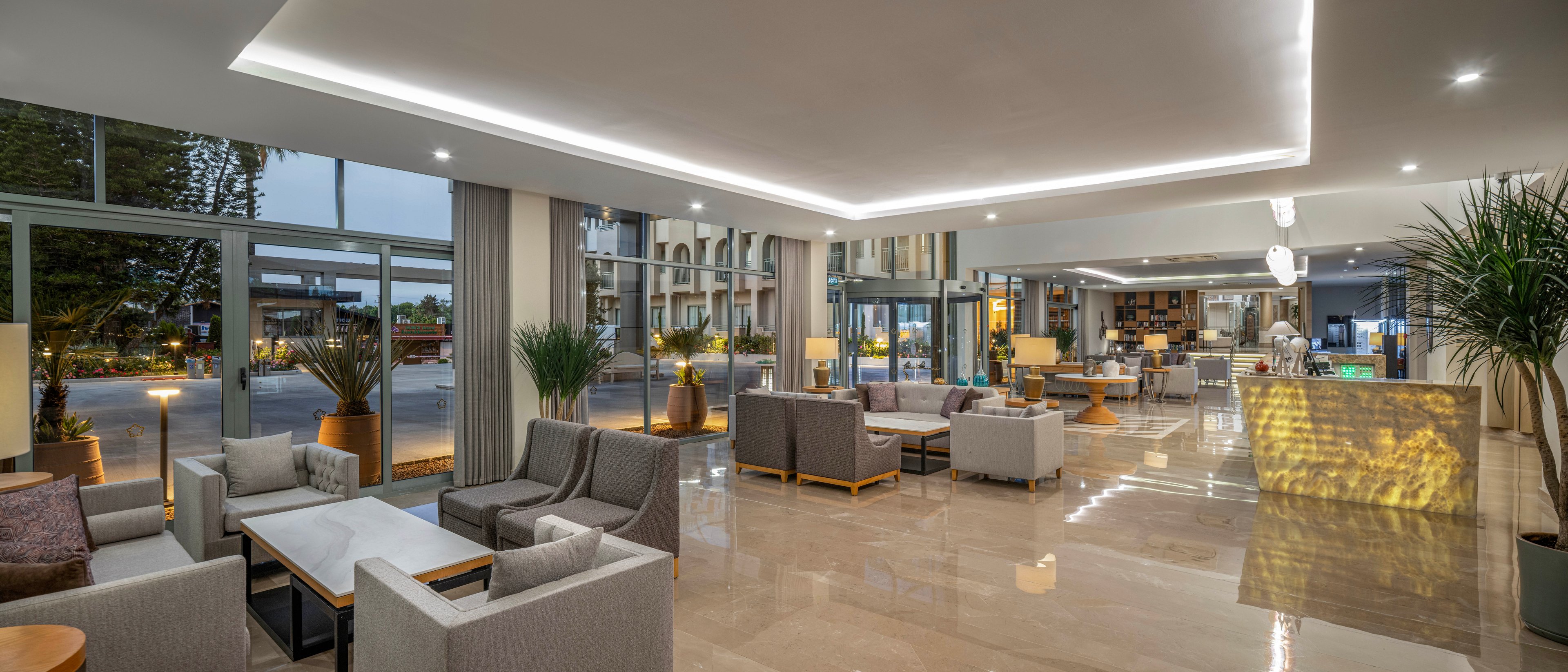 A contemporary hotel lobby features multiple seating areas with armchairs and sofas arranged around low tables. Large floor-to-ceiling windows reveal an exterior street and trees outside. Warm ambient lighting from floor lamps and recessed ceiling lights reflects on the polished tile floor. A reception desk and decorative plants add to the welcoming, upscale atmosphere.
