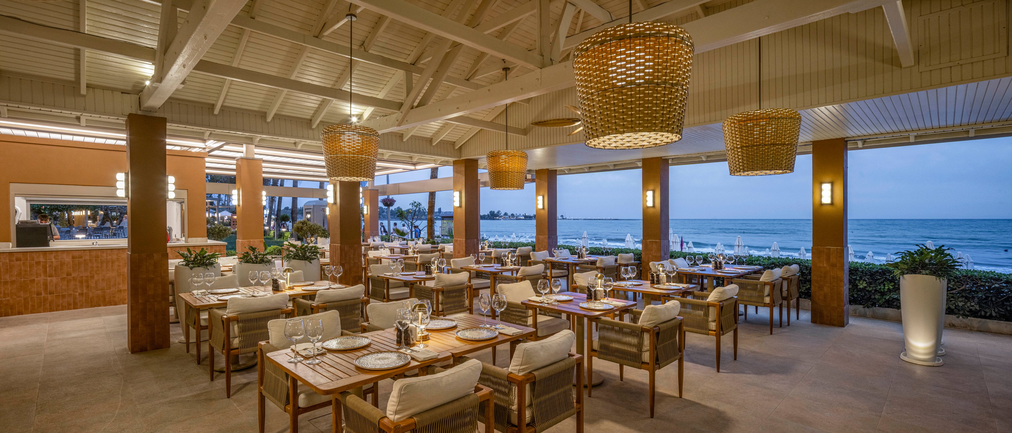An elegant open-air restaurant overlooks the ocean at dusk, with neatly arranged wooden tables and cushioned chairs. Warm pendant lights and wall sconces create a cozy glow under the high white-beamed ceiling. The setting features a relaxed coastal ambiance with views of the shoreline and calm water in the background. Table settings and soft lighting suggest a refined yet casual dining experience by the sea.
