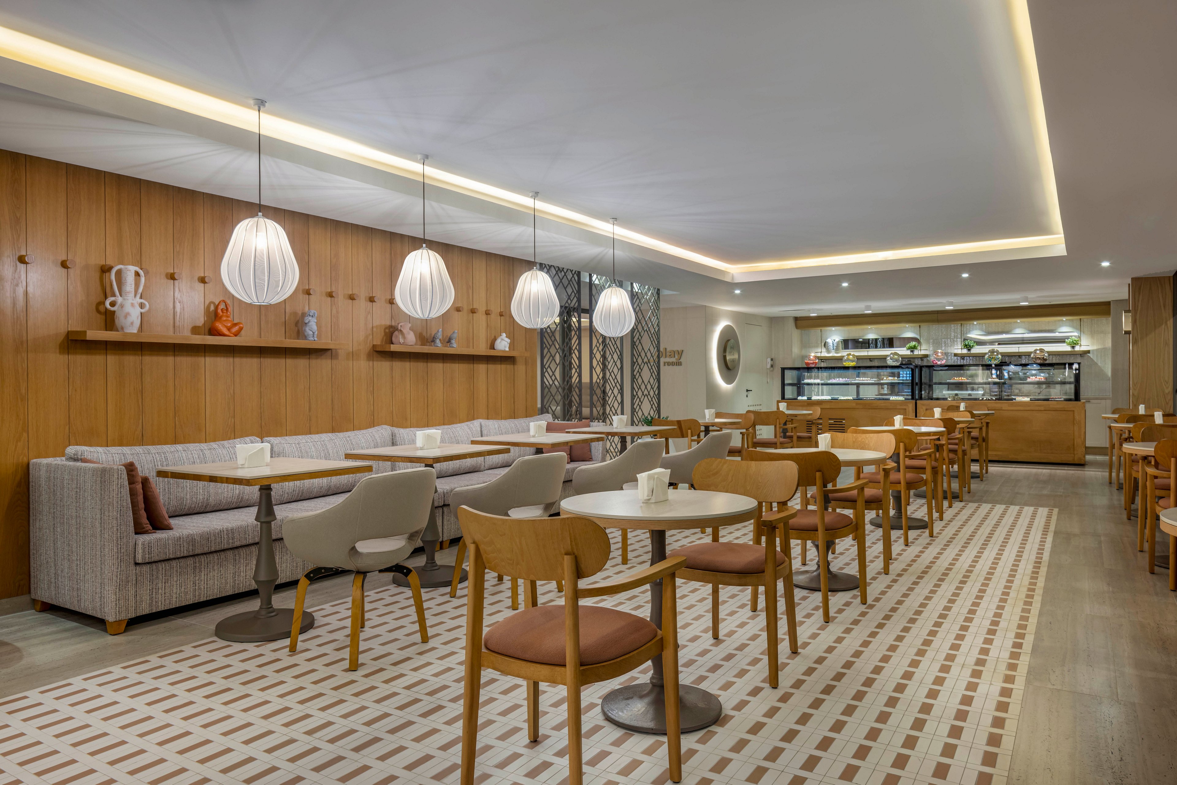 A contemporary café interior features neatly arranged round tables and wooden chairs in a bright, inviting space. Soft pendant lights hang above a long upholstered bench along a wood-paneled wall. In the background, a display counter and shelves showcase food and beverages. The neutral color palette and clean lines create a calm, minimalist atmosphere suitable for dining or casual meetings.