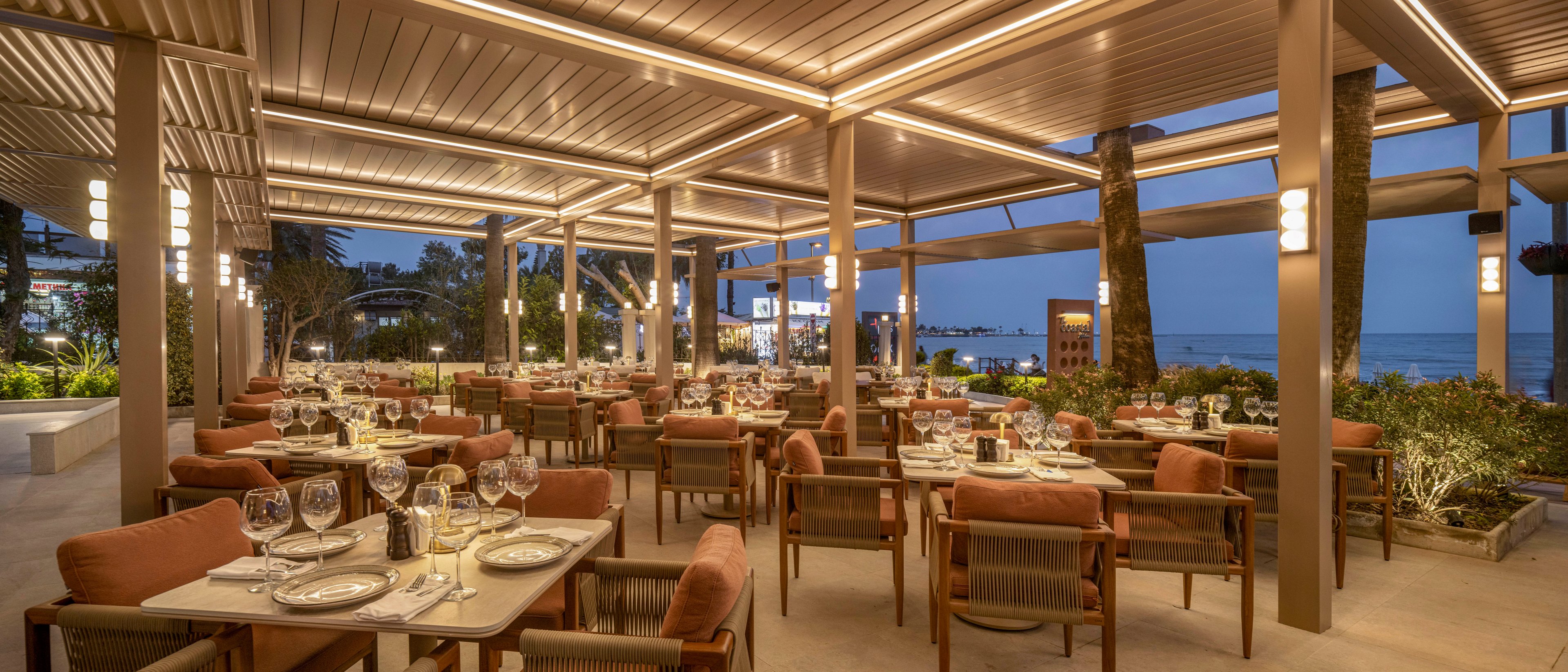 An upscale open-air restaurant terrace is set for dinner with neatly arranged tables and cushioned chairs. Warm ambient lighting from the ceiling and lantern-style fixtures creates a cozy atmosphere as evening falls. Glassware, plates, and cutlery are carefully placed, suggesting a fine dining experience. In the background, palm trees and a glimpse of the sea enhance the relaxed, coastal setting.