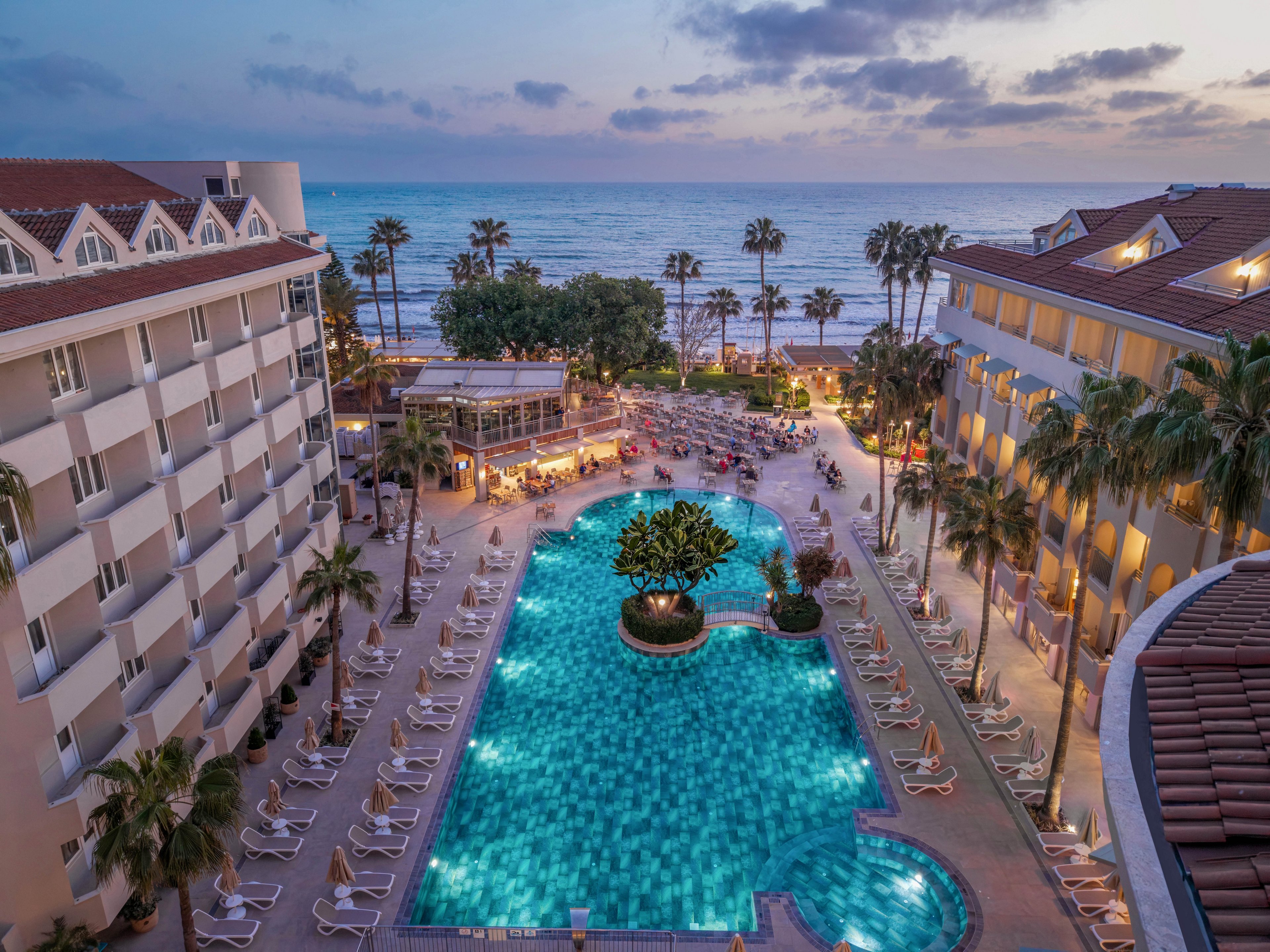 An elevated view overlooks a large resort swimming pool surrounded by lounge chairs and palm trees. Guests gather around the pool and outdoor dining areas as evening lights begin to glow. The scene opens toward a sandy beach and calm ocean under a colorful sunset sky. Warm building lights and reflections in the water create a relaxed, vacation atmosphere.