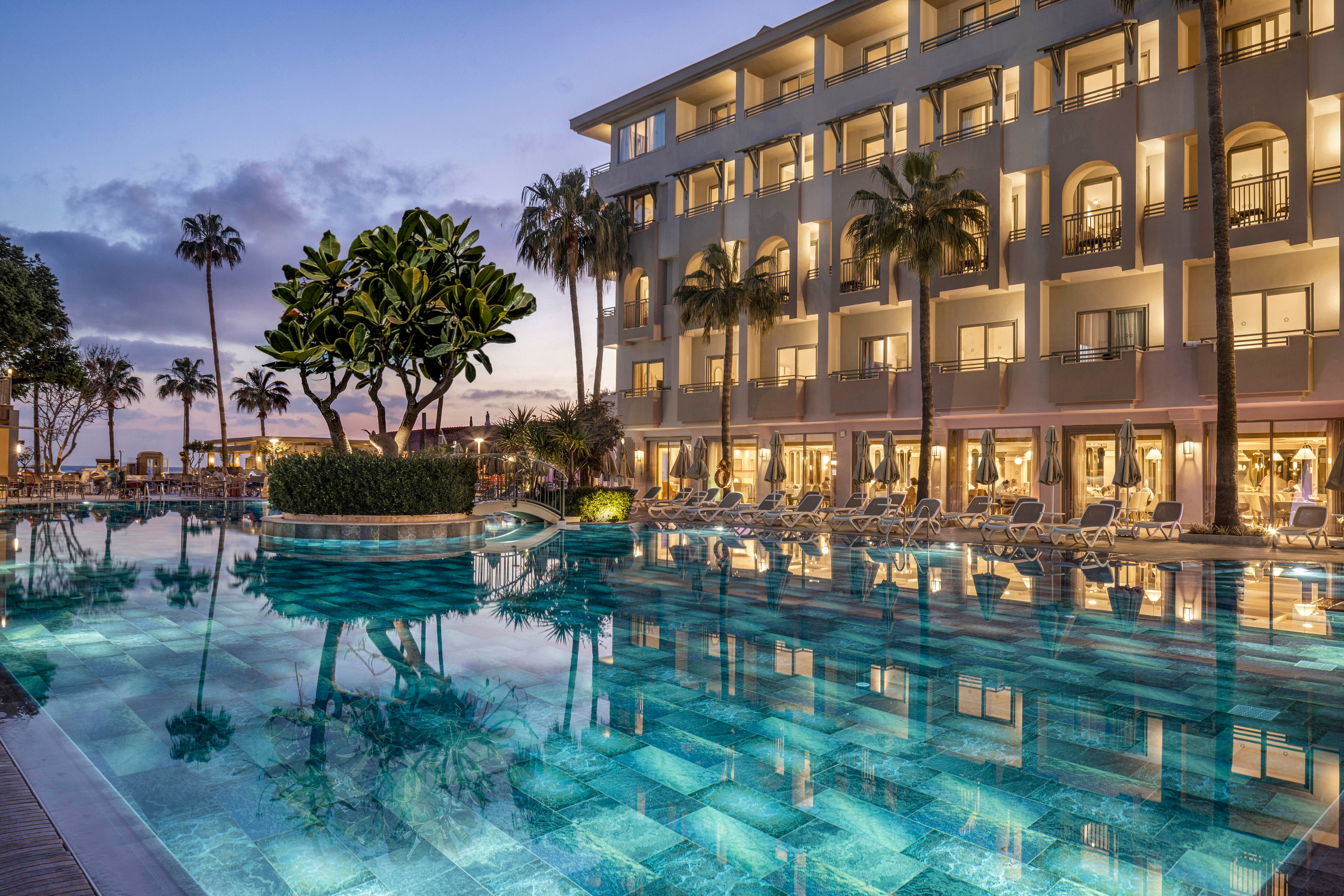 A luxurious resort pool is shown at dusk, reflecting the warm lights of a multi-story hotel building. Palm trees and lush greenery surround the pool, creating a tropical atmosphere. Lounge chairs line the poolside, suggesting a relaxing vacation setting. The sky transitions from soft blue to purple tones, enhancing the tranquil and elegant mood of the scene.