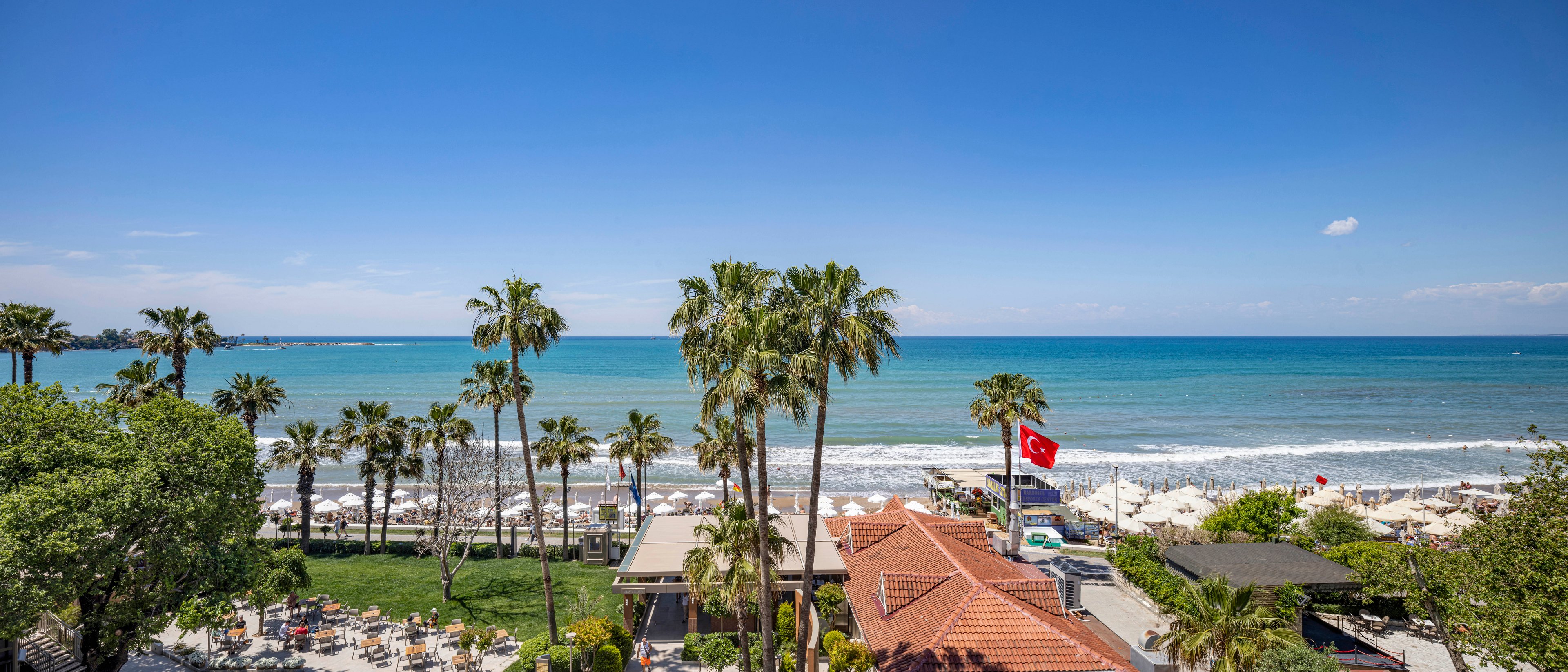 A coastal resort overlooks a bright blue sea under a clear sky. Tall palm trees line the beachfront, separating a green lawn from rows of sun loungers and umbrellas on the sand. A red national flag waves above a building with a terracotta roof near the shoreline. The calm waves and scattered beachgoers create a relaxed holiday atmosphere.