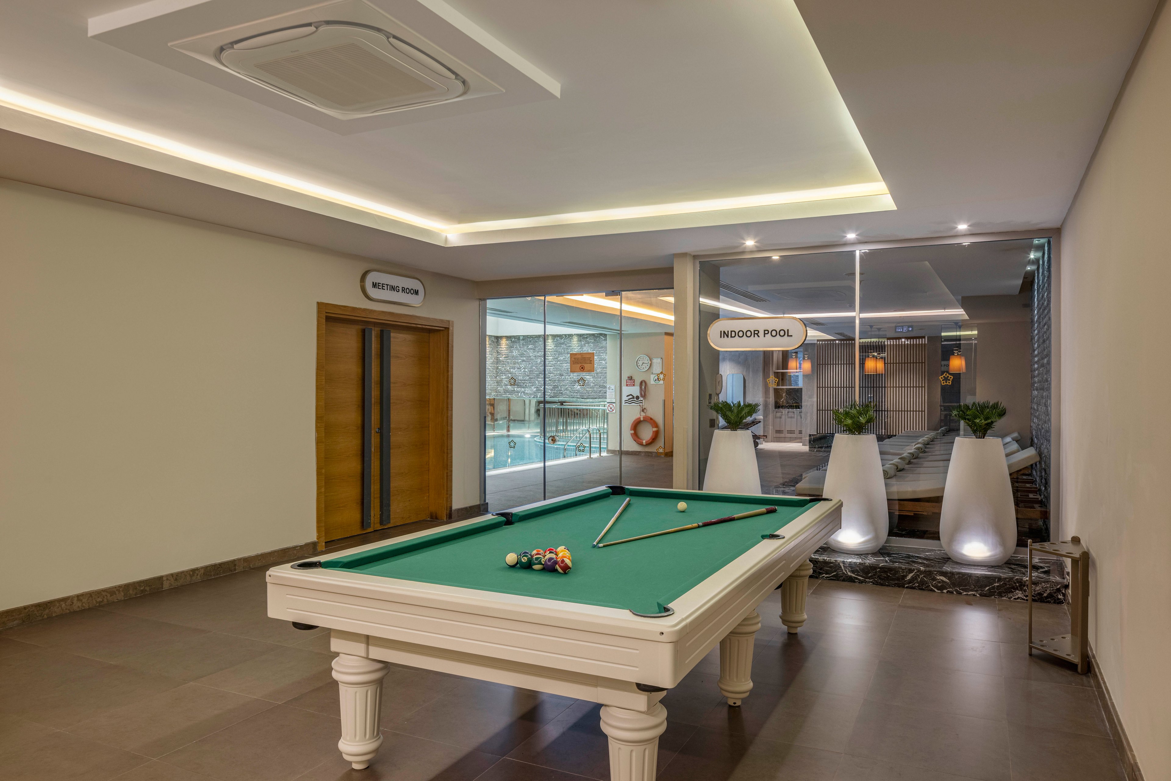 A white billiards table with balls and cues stands in a modern indoor recreation area. Behind it, glass walls reveal an illuminated indoor pool with lounge chairs and decorative lighting. Signs reading "Meeting Room" and "Indoor Pool" are visible above the doors and entrance. The space features neutral tones, recessed ceiling lights, and large white planters with green plants, creating a clean and contemporary atmosphere.