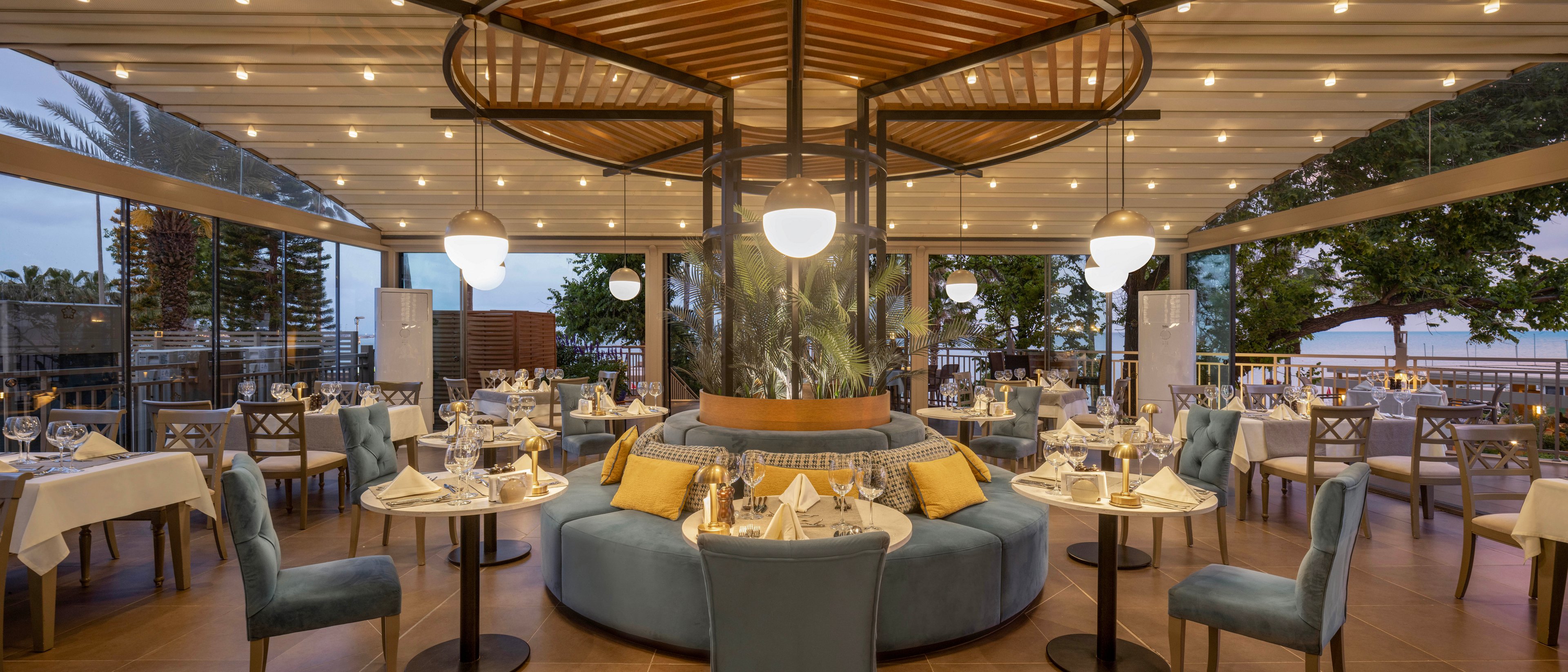 A sophisticated restaurant interior features a central circular banquette surrounded by neatly arranged dining tables. Soft blue chairs and cushioned seating create a comfortable and upscale atmosphere. Warm string lights and hanging globe fixtures illuminate the space, while large windows reveal palm trees and a coastal view at dusk. The table settings with glassware and folded napkins suggest a fine dining experience in a relaxed seaside setting.