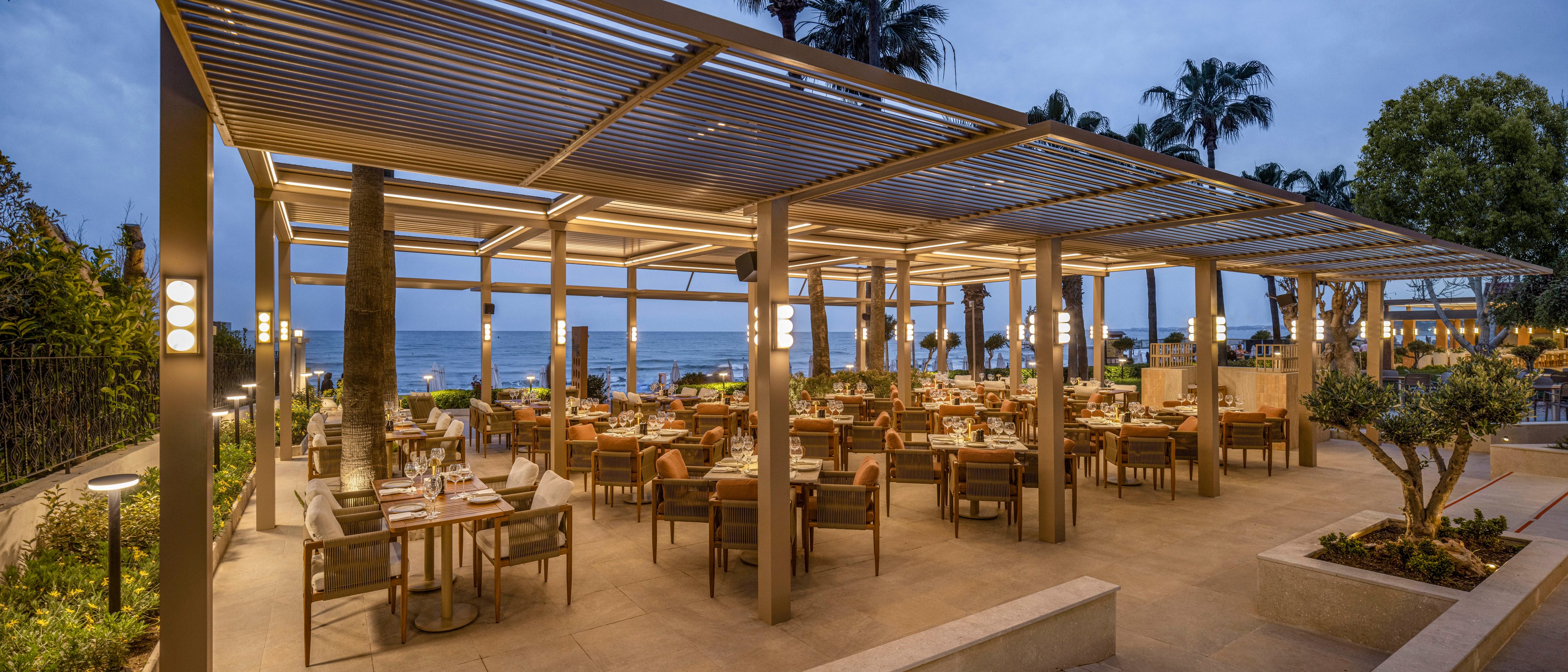 An elegant open-air restaurant is set under modern pergolas near the sea at dusk. Neatly arranged wooden tables and chairs are prepared for guests with place settings and soft lighting. Warm lamps and subtle architectural lines create a cozy yet contemporary atmosphere. Palm trees and the ocean are visible in the background, enhancing the relaxed coastal setting.