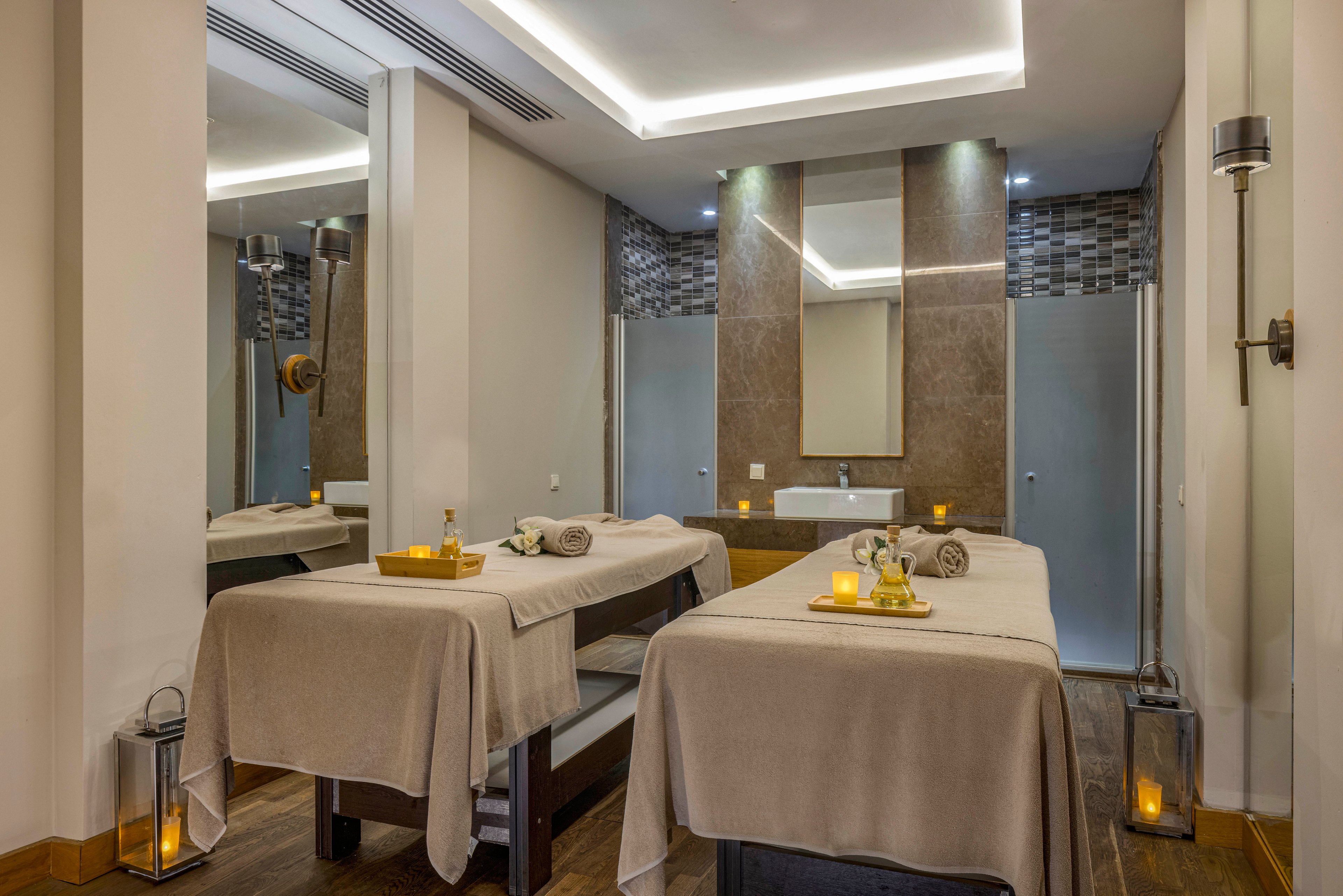 A serene spa treatment room features two massage tables neatly prepared with towels and small trays of oils. Soft lighting from recessed ceiling lights and glowing candles creates a warm, relaxing atmosphere. A large mirror and modern wall panels frame the sink area at the back of the room. Lanterns and minimalist decor enhance the tranquil, upscale spa setting.