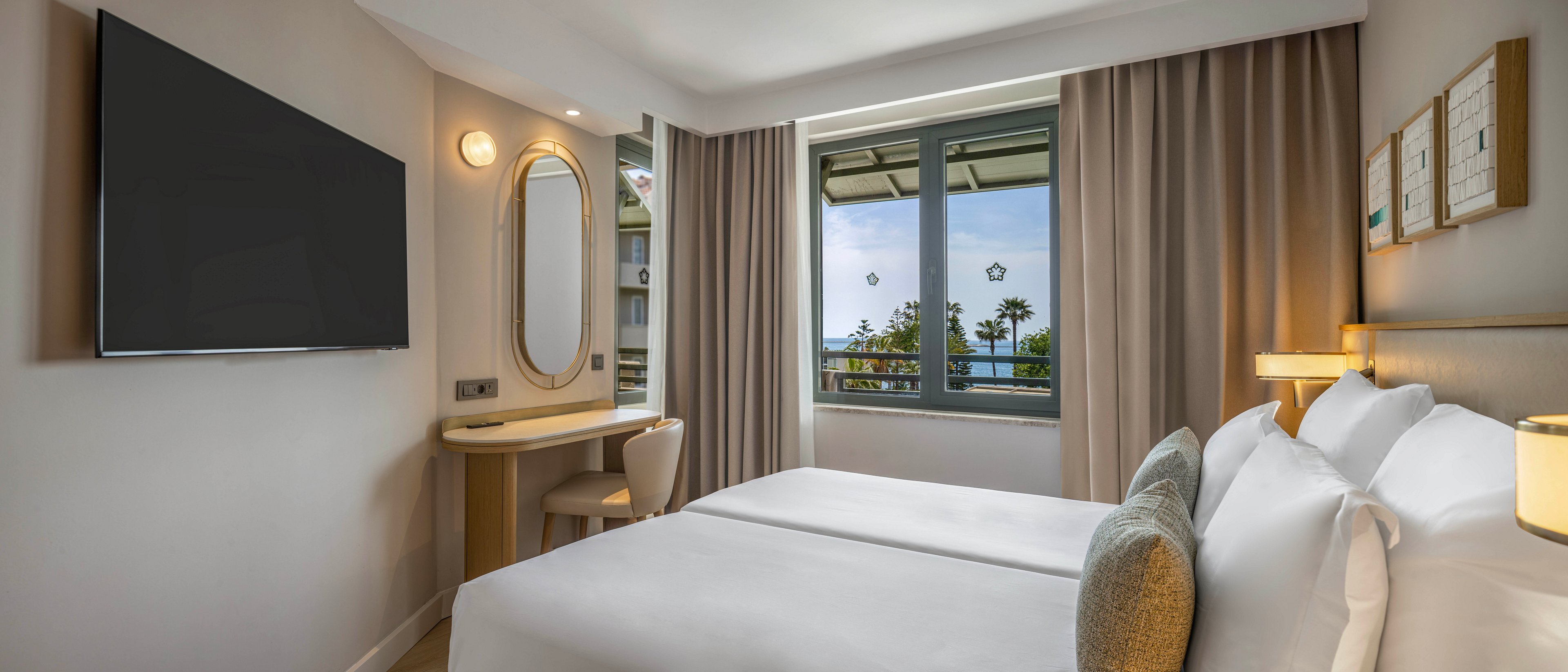 A contemporary hotel room features two neatly made twin beds with white linens and neutral-toned cushions. A large flat-screen television is mounted on the wall opposite the beds, and a compact vanity desk with an oval mirror sits beside it. Floor-to-ceiling curtains frame a wide window that reveals a view of palm trees and the sea outside. Soft lighting and beige walls create a calm, inviting atmosphere suitable for relaxation or business travel.