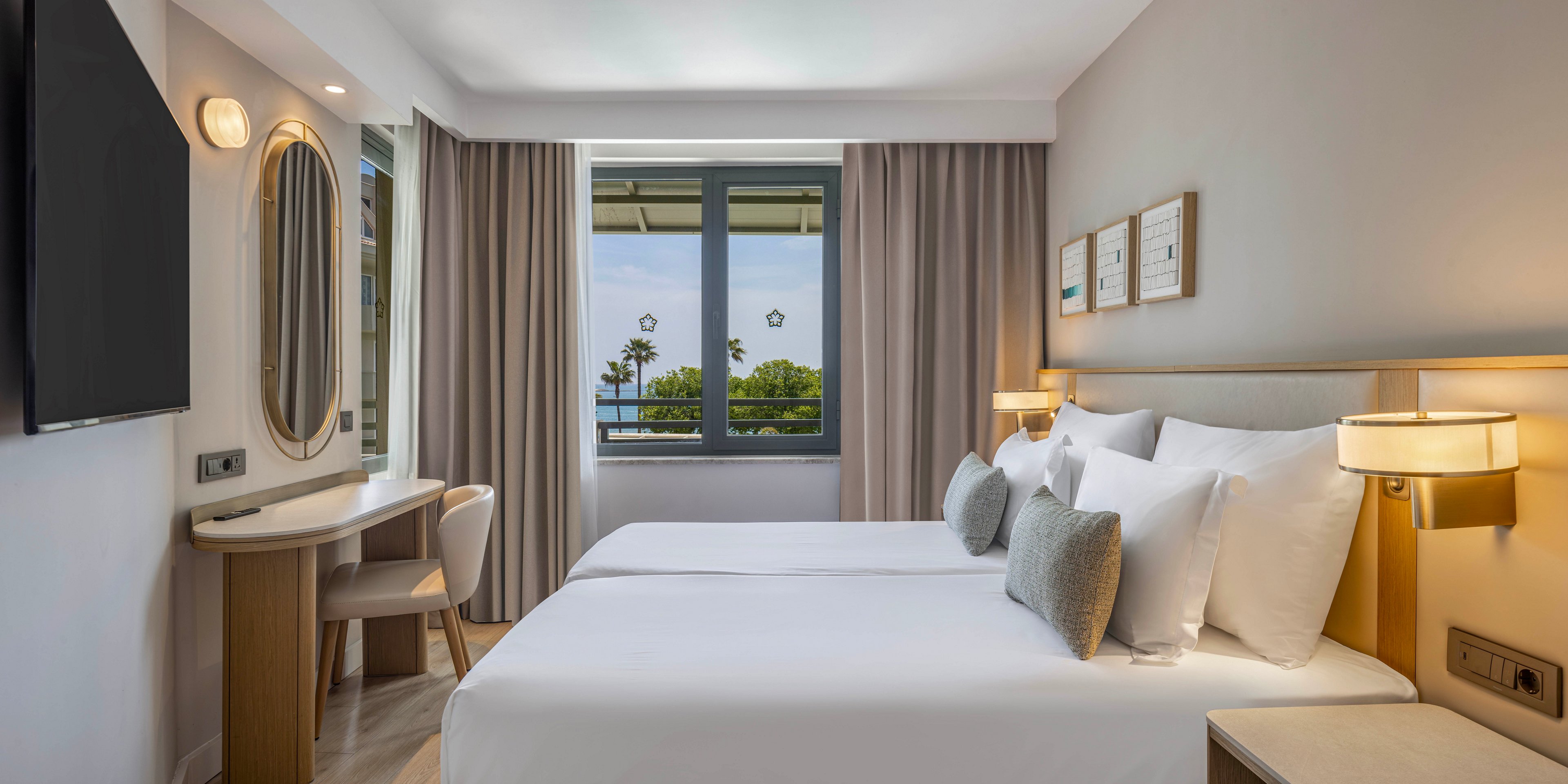 A contemporary hotel room features two neatly made twin beds with white linens and gray accent pillows. The space includes a wall-mounted flat-screen TV, a wooden desk with a round mirror, and soft ambient lighting from wall lamps. Large curtains frame a central window that offers a view of trees and a distant body of water. Neutral tones and clean lines create a calm, minimalist atmosphere suitable for relaxation or business travel.
