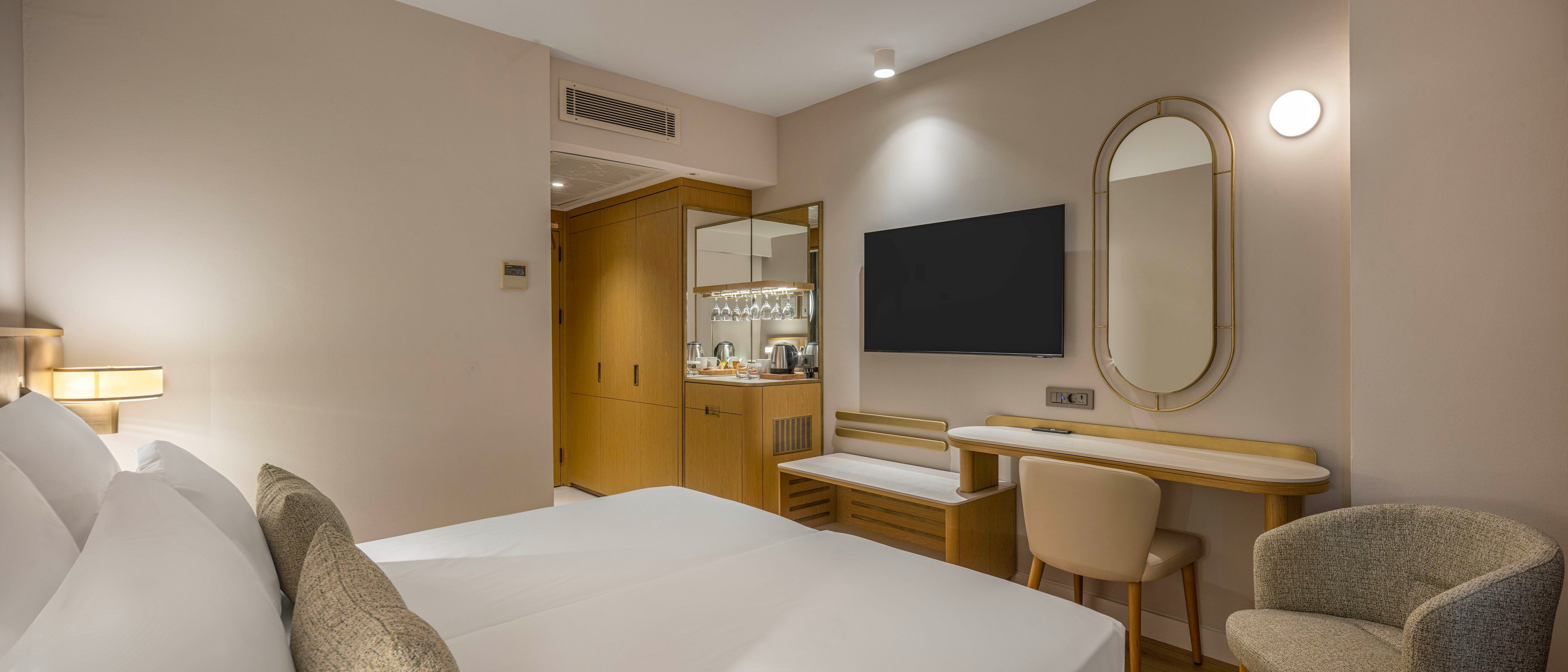A contemporary hotel room features a neatly made double bed with white linens in the foreground. The space includes a wall-mounted flat-screen television, a minimalist desk with a round mirror, and a cushioned chair. Warm wood cabinetry houses a small minibar area with glassware and a mirror backsplash. Soft, neutral tones and subtle lighting create a calm and inviting atmosphere suitable for relaxation or business travel.