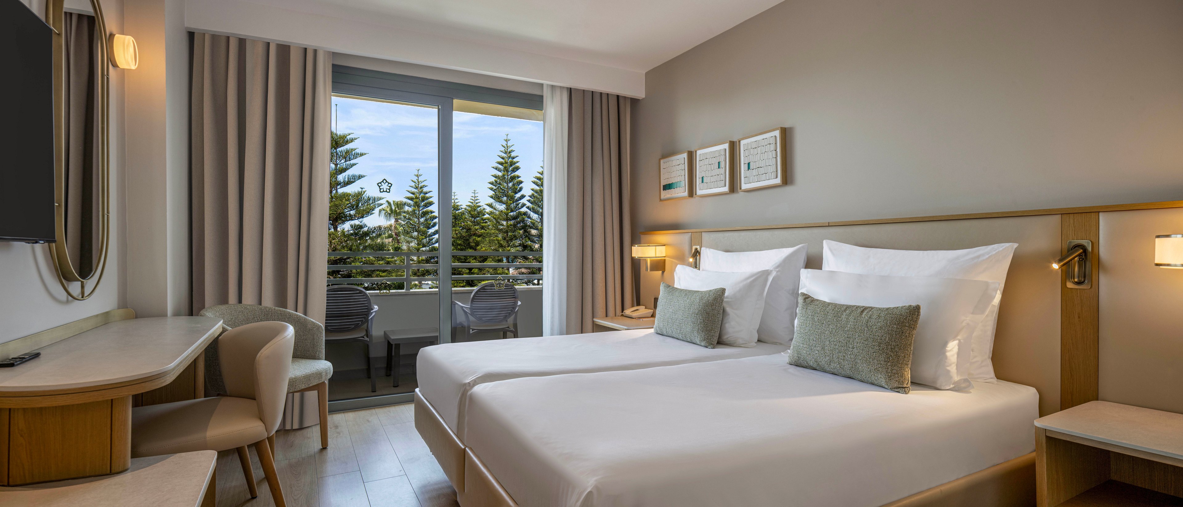 A contemporary hotel room features two neatly made twin beds with white linens and soft green accent pillows. The space includes a wooden desk with a chair, a wall-mounted television, and bedside tables with warm lamps. Large curtains frame a glass door leading to a balcony with outdoor chairs and a view of evergreen trees. Neutral tones and soft lighting create a calm, inviting atmosphere ideal for relaxation or business travel.