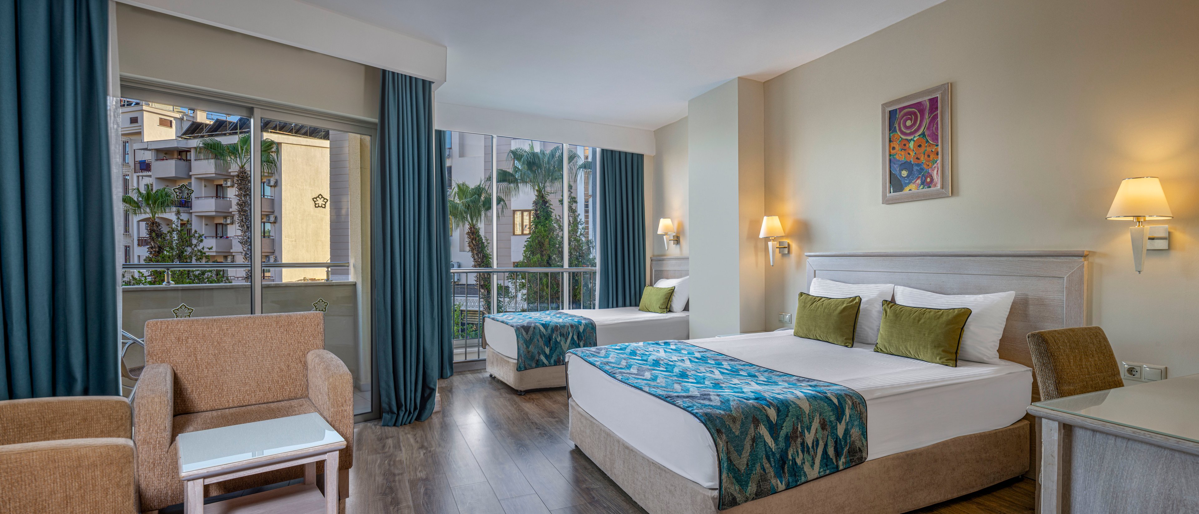 Bright contemporary hotel room featuring two neatly made twin beds with patterned blue runners and green accent pillows. The space includes a small seating area with armchairs and a side table, set on dark wood flooring. Large windows and a glass balcony door reveal an exterior view of palm trees and nearby buildings. Soft lighting from wall sconces and a colorful framed artwork create a calm, welcoming atmosphere.