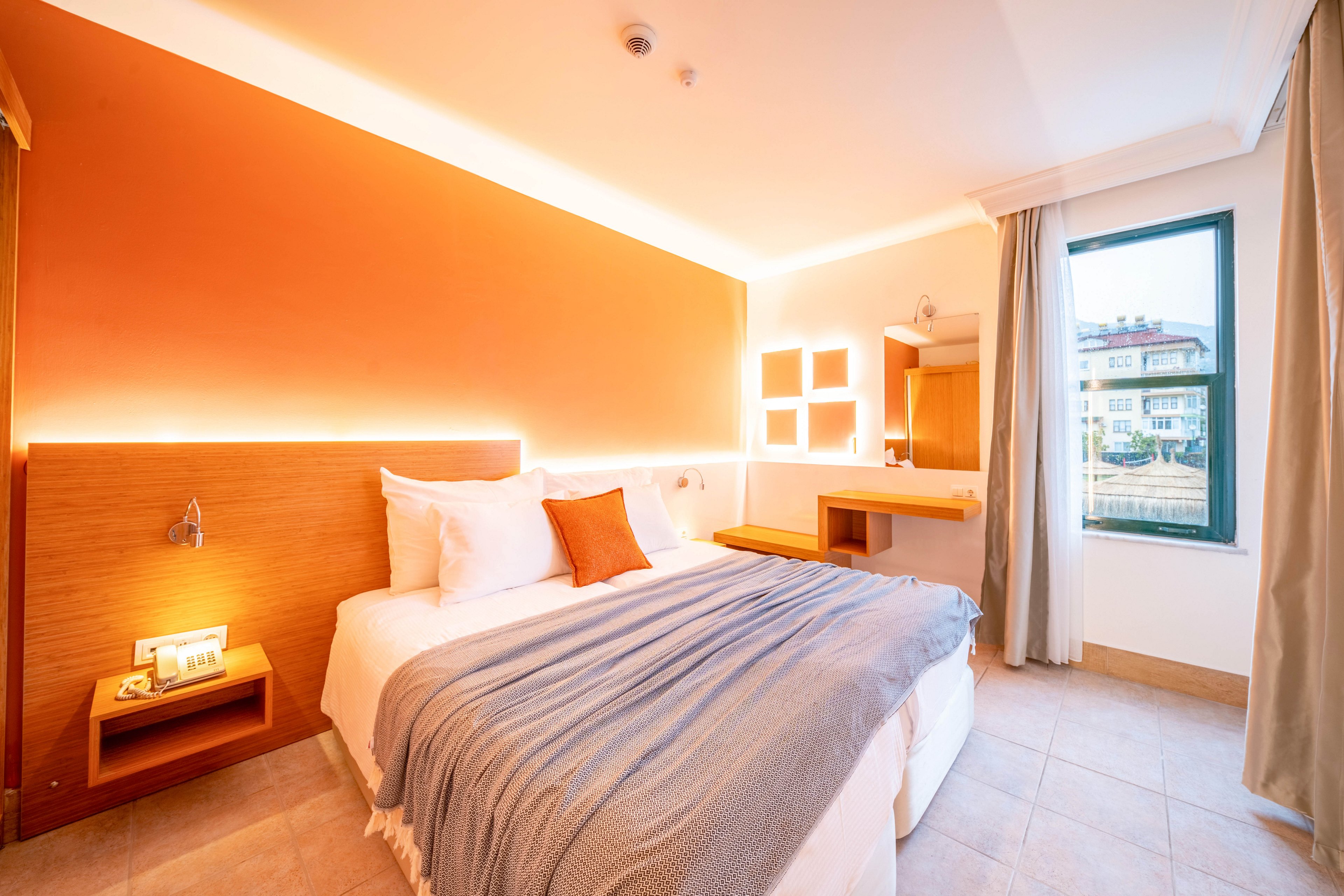 A contemporary hotel bedroom features a neatly made double bed with white linens, a gray throw blanket, and an orange accent pillow. The room is illuminated by warm indirect lighting along an orange accent wall and above the headboard. A small bedside table, wall-mounted reading light, and minimalist desk area with mirror complete the interior. Natural light enters through a large window with curtains, revealing buildings outside.